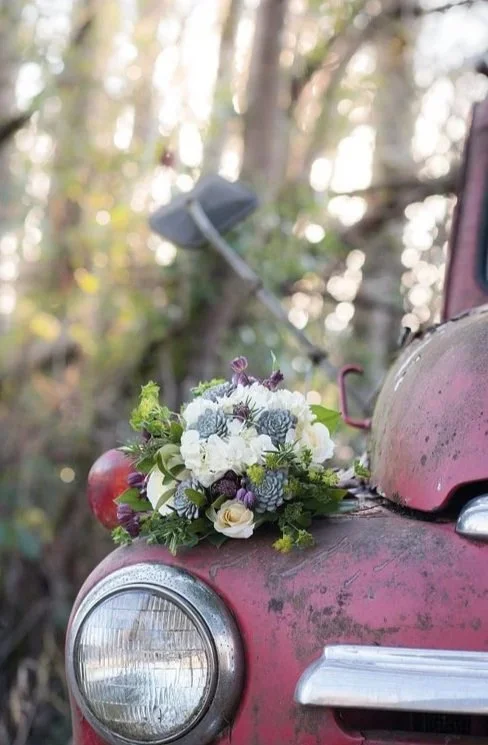 A bouquet of white, purple, and green flowers resting on the hood of a vintage red car with a slightly rusted exterior, outdoors in a wooded area.