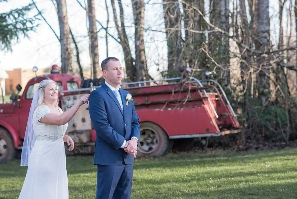 Bride in a white dress touching the groom’s shoulder outside during a wedding ceremony with an old red fire truck and trees in the background.
