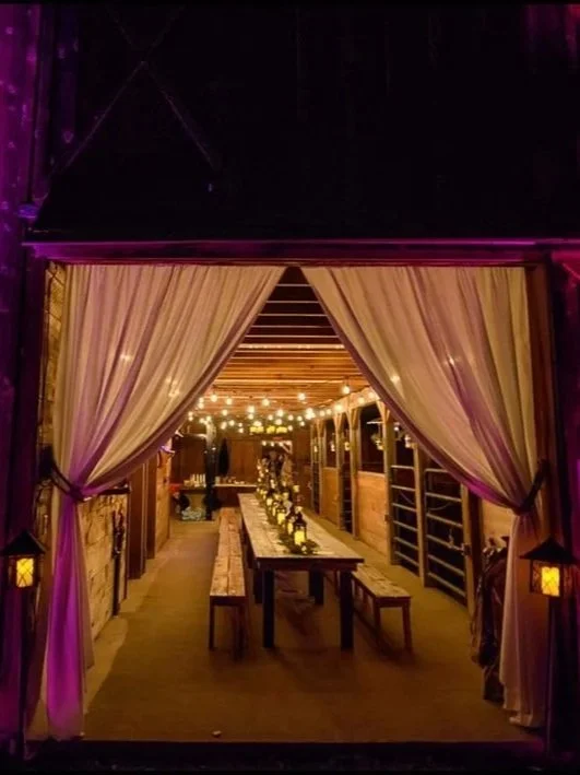 Decorated rustic indoor venue with a central long table adorned with candle lanterns, illuminated by string lights, seen through open curtains