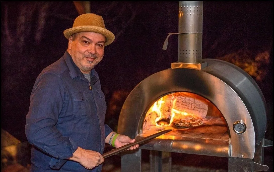 Man wearing a hat and blue shirt using a peel remover in front of a pizza oven with flames