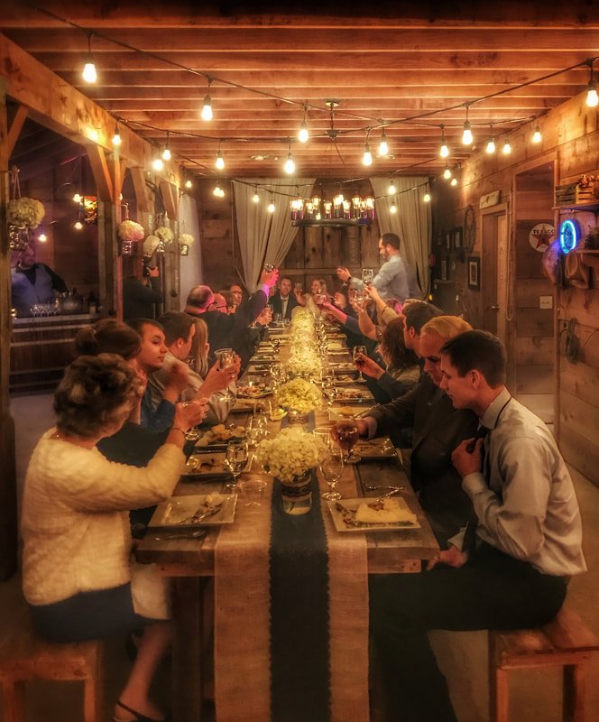 People gathered around a long table in a rustic decorated wooden dining room for a celebration or dinner event, with string lights hanging from the ceiling.