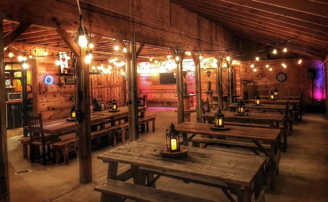 Interior of a rustic restaurant or bar with wooden tables and benches, string lights hanging from the ceiling, candles on the tables, and a dartboard on the wall.