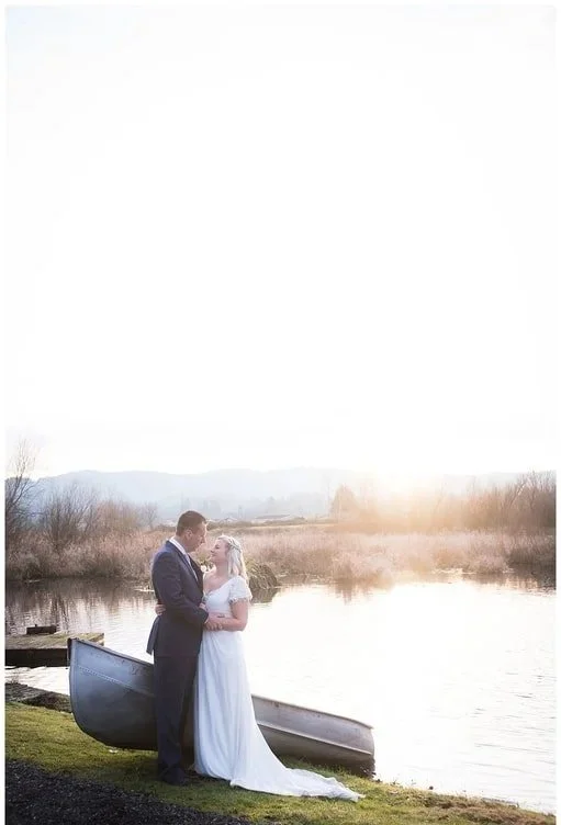 A bride and groom stand by a lake during sunset, embracing each other with a canoe nearby.