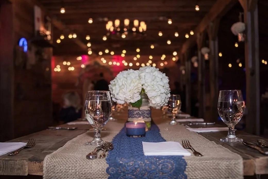 Wedding reception table with a flower centerpiece, glassware, silverware, and a burlap table runner in a rustic indoor setting with string lights.