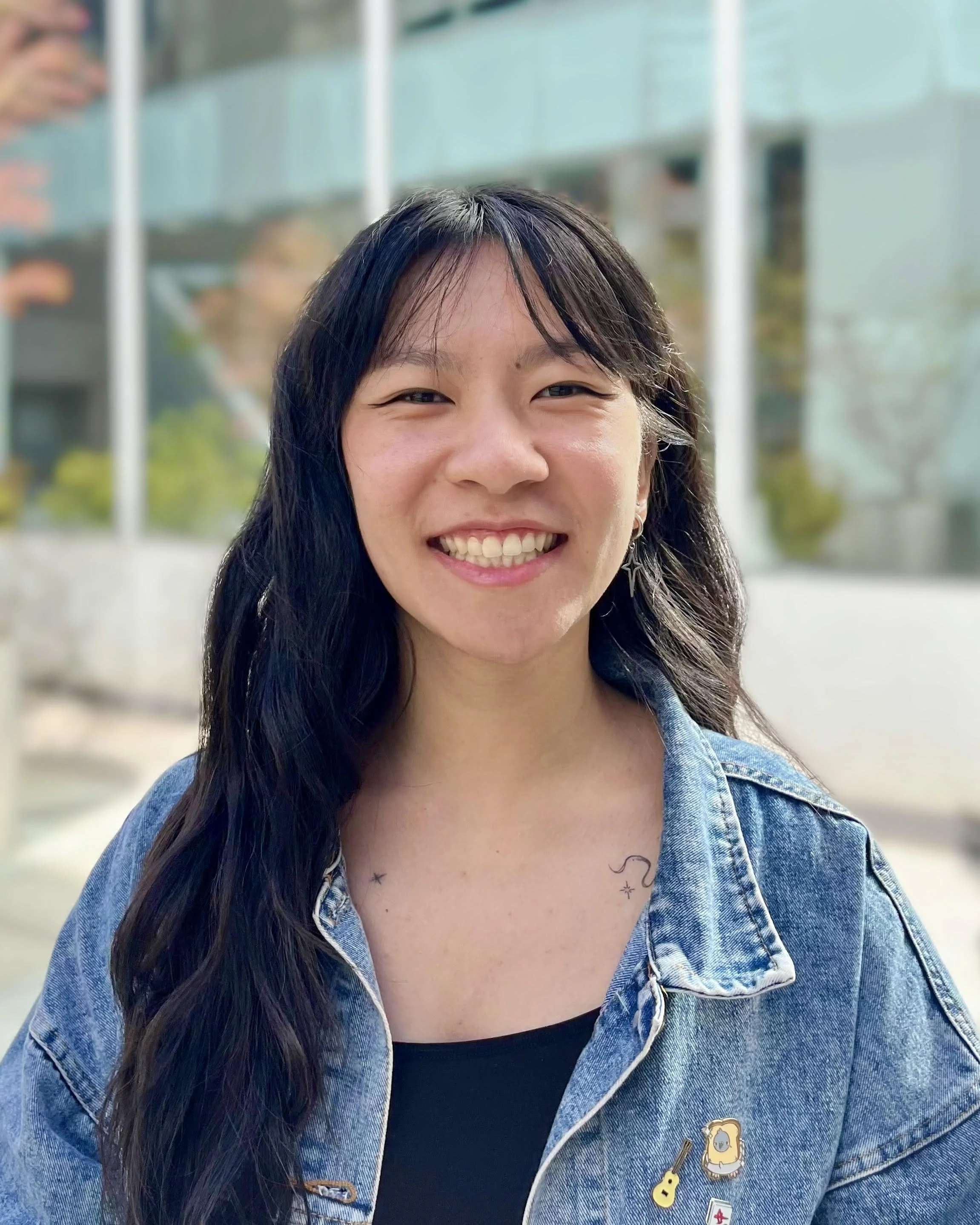 A person with long wavy black hair, smiling outdoors, wearing a denim jacket with pins and a black top, with a glass building and trees in the background.