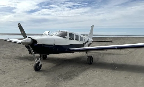 Small white and black single-engine airplane on a sandy runway with cloudy sky in the background.