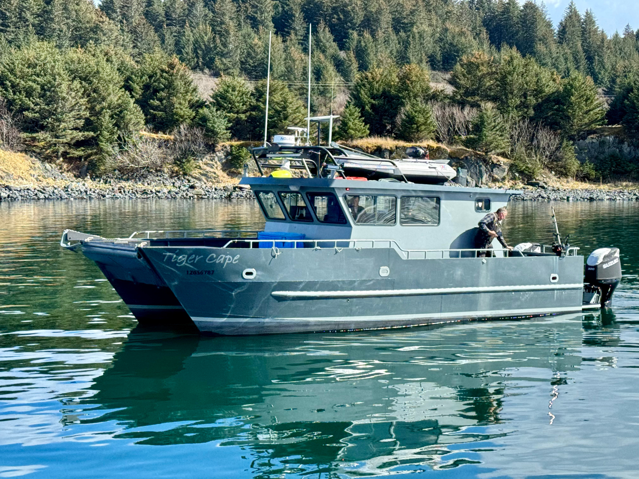 A boat named 'Tiger Cape' with a person standing at the stern, floating on calm water near a forested shoreline with rocks and trees.