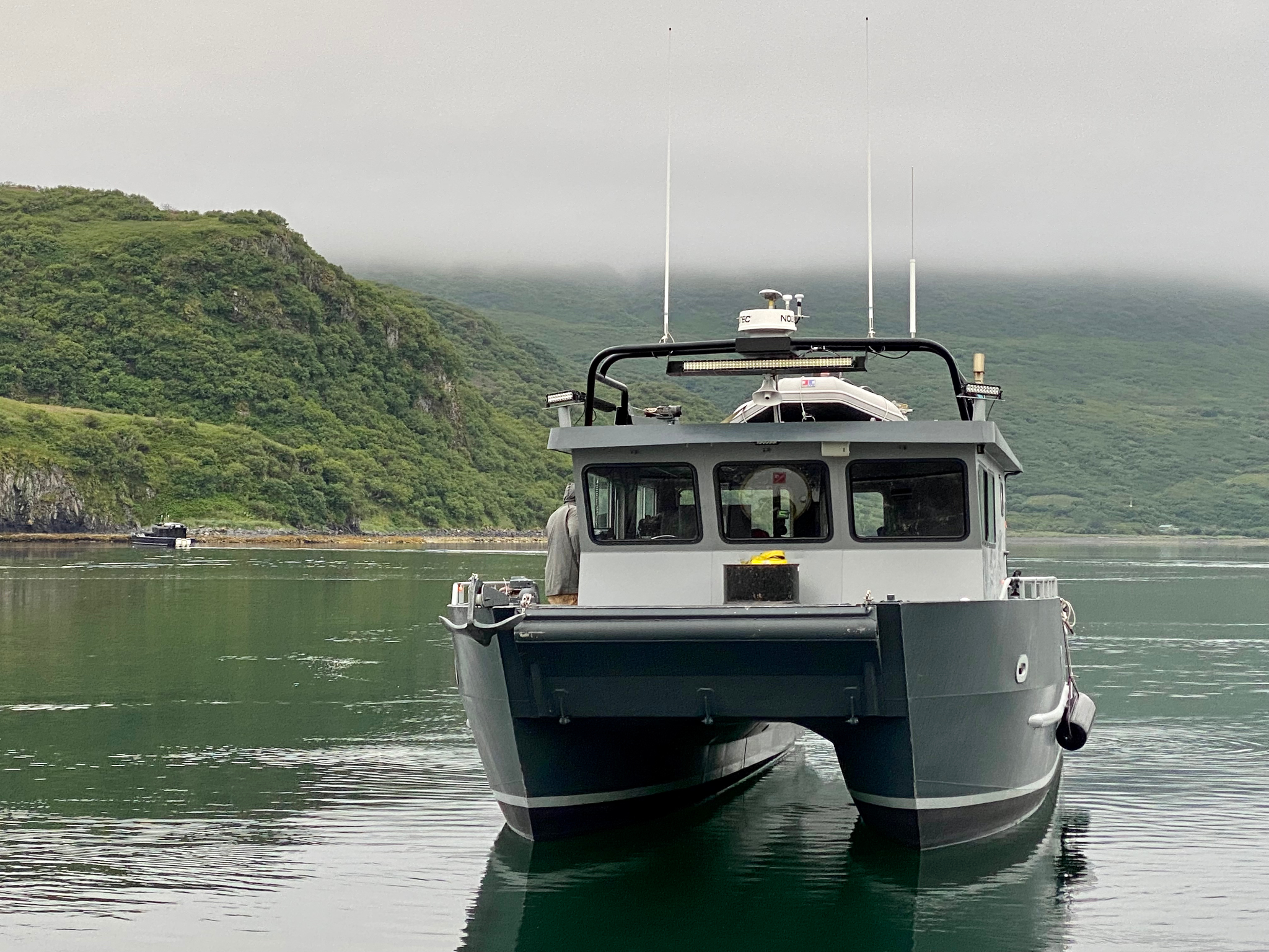 A boat on calm water with a green, hilly landscape in the background, partially obscured by fog.