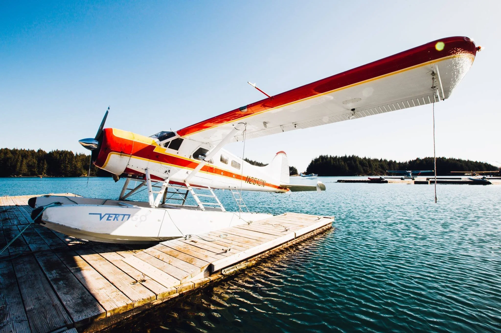 A seaplane with red, yellow, and white colors docked at a wooden pier on a body of water with trees in the background under a clear blue sky.
