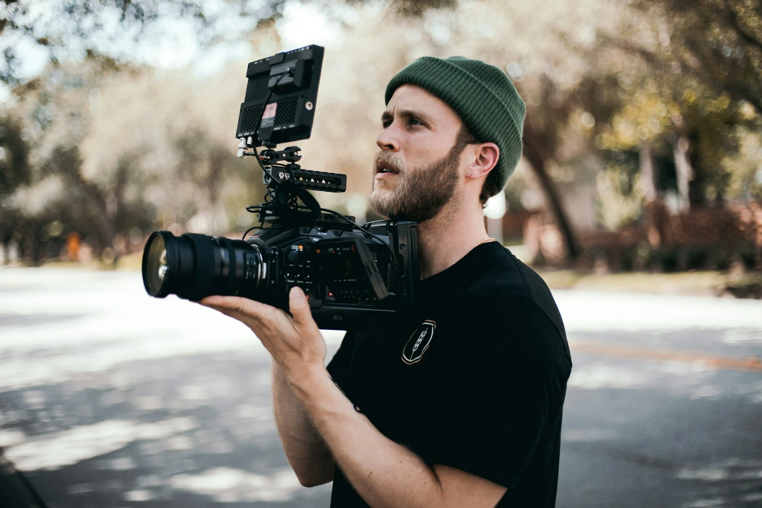 A man with a beard and green beanie is holding a professional video camera with an external monitor attached, outdoors with trees and a blurred background.