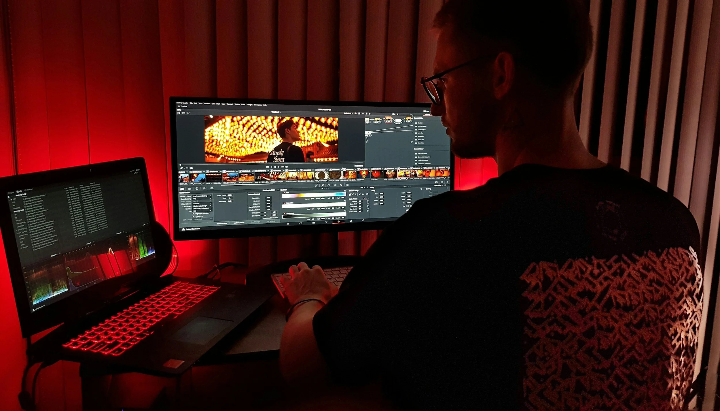 A person working on video editing at a desk with two monitors in a dimly lit room with red lighting and wooden slatted wall.