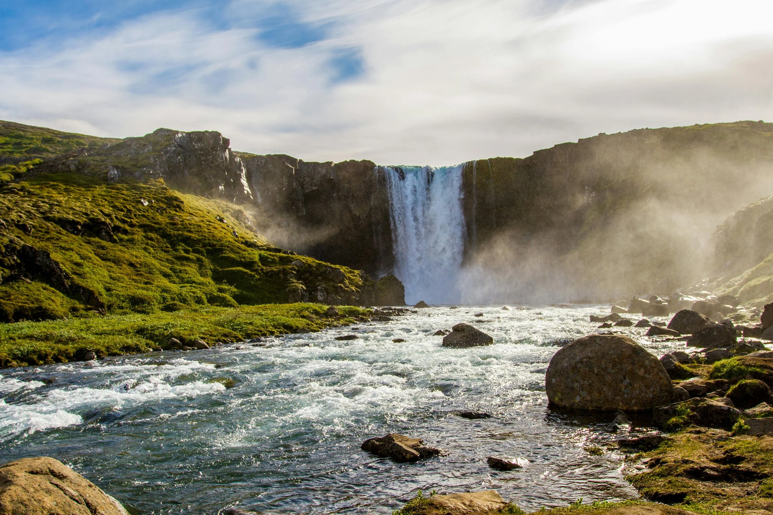 Wasserfall in einer grünen Landschaft mit fließendem Fluss im Vordergrund, teilweise bewölkter Himmel.