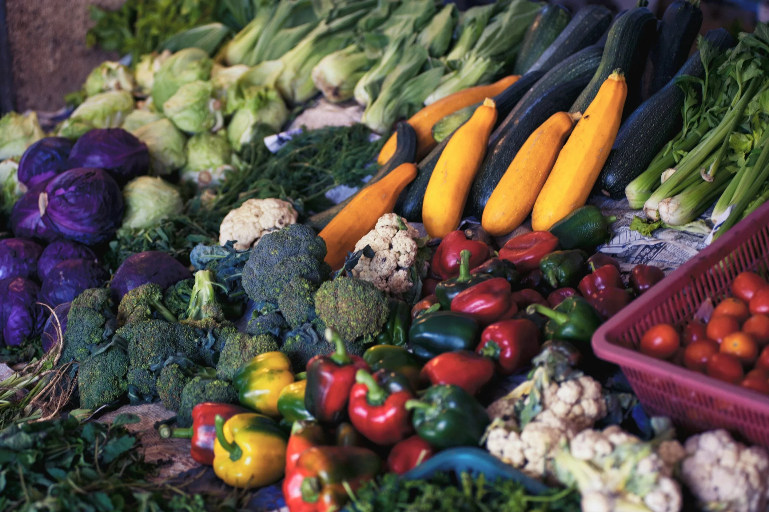 A display of fresh vegetables including purple eggplants, cauliflower, broccoli, yellow squash, zucchini, peppers, and tomatoes at a farmers market.