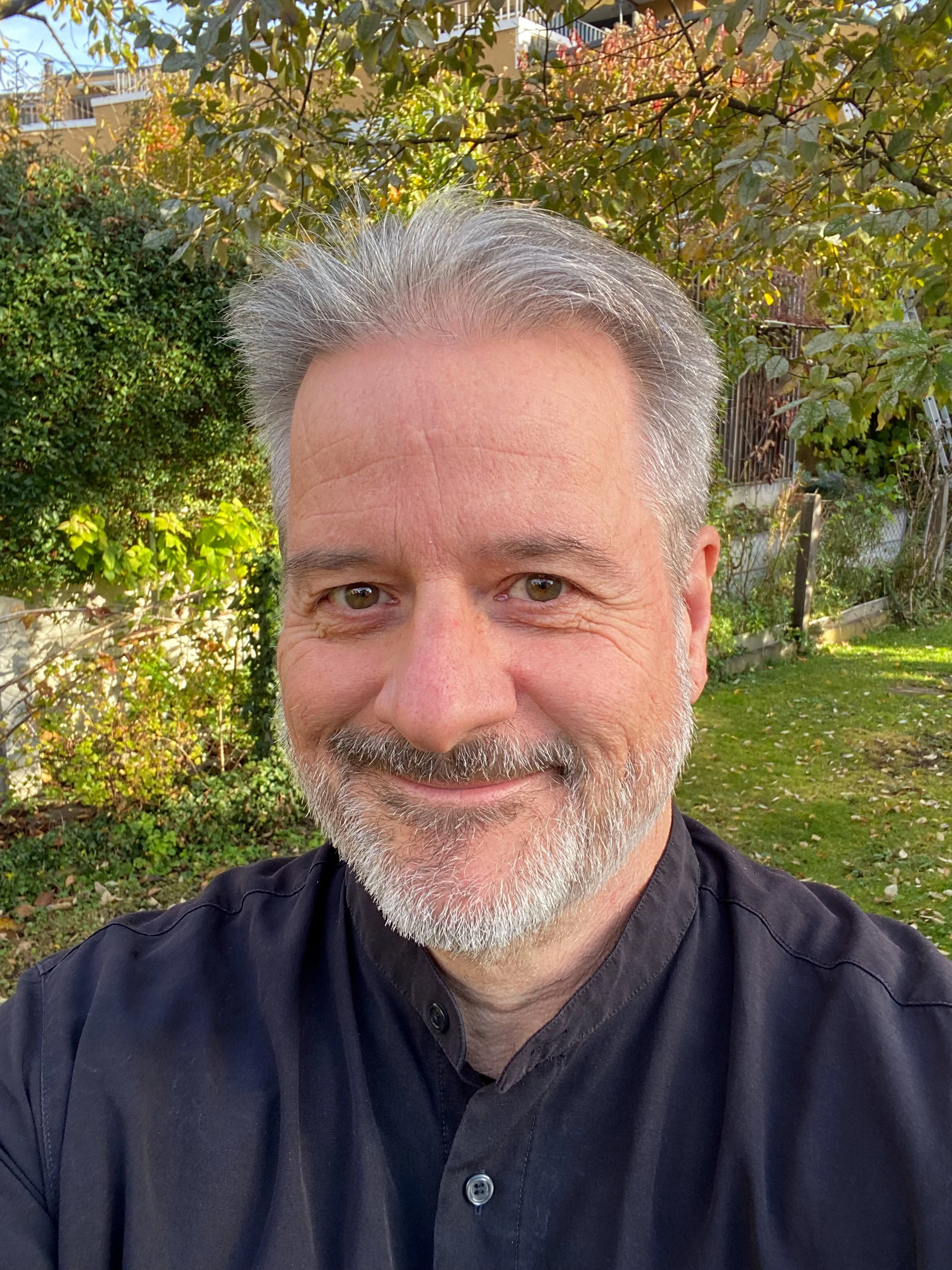 A smiling middle-aged man with gray hair and a beard taking a selfie outdoors in a garden with trees and shrubs in the background.