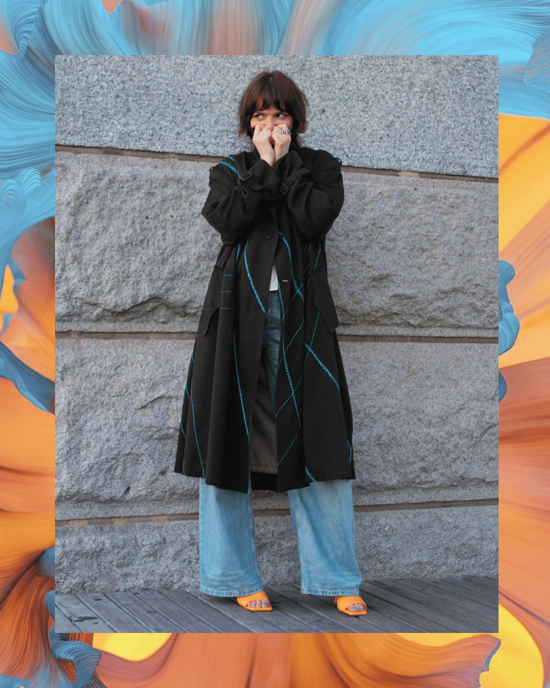 A woman standing in front of a stone wall, dressed in a long black coat with blue stitching, wide-leg jeans, and yellow shoes, with her hands near her face and looking to the side.