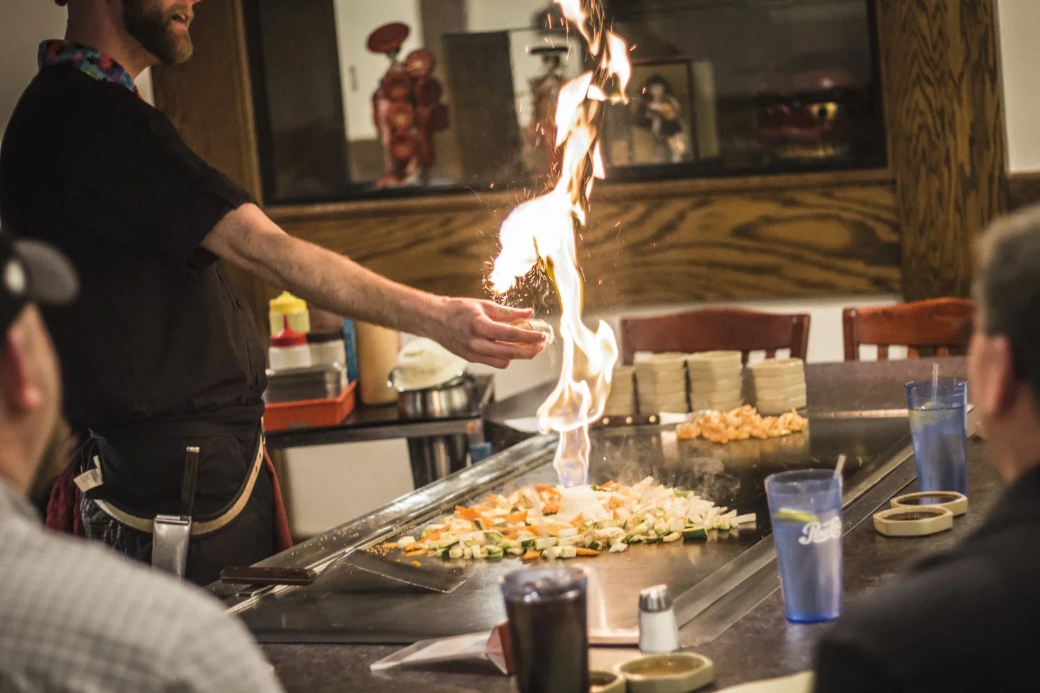 A chef performs a flaming cooking technique at a teppanyaki restaurant, with vegetables on the grill and patrons watching.