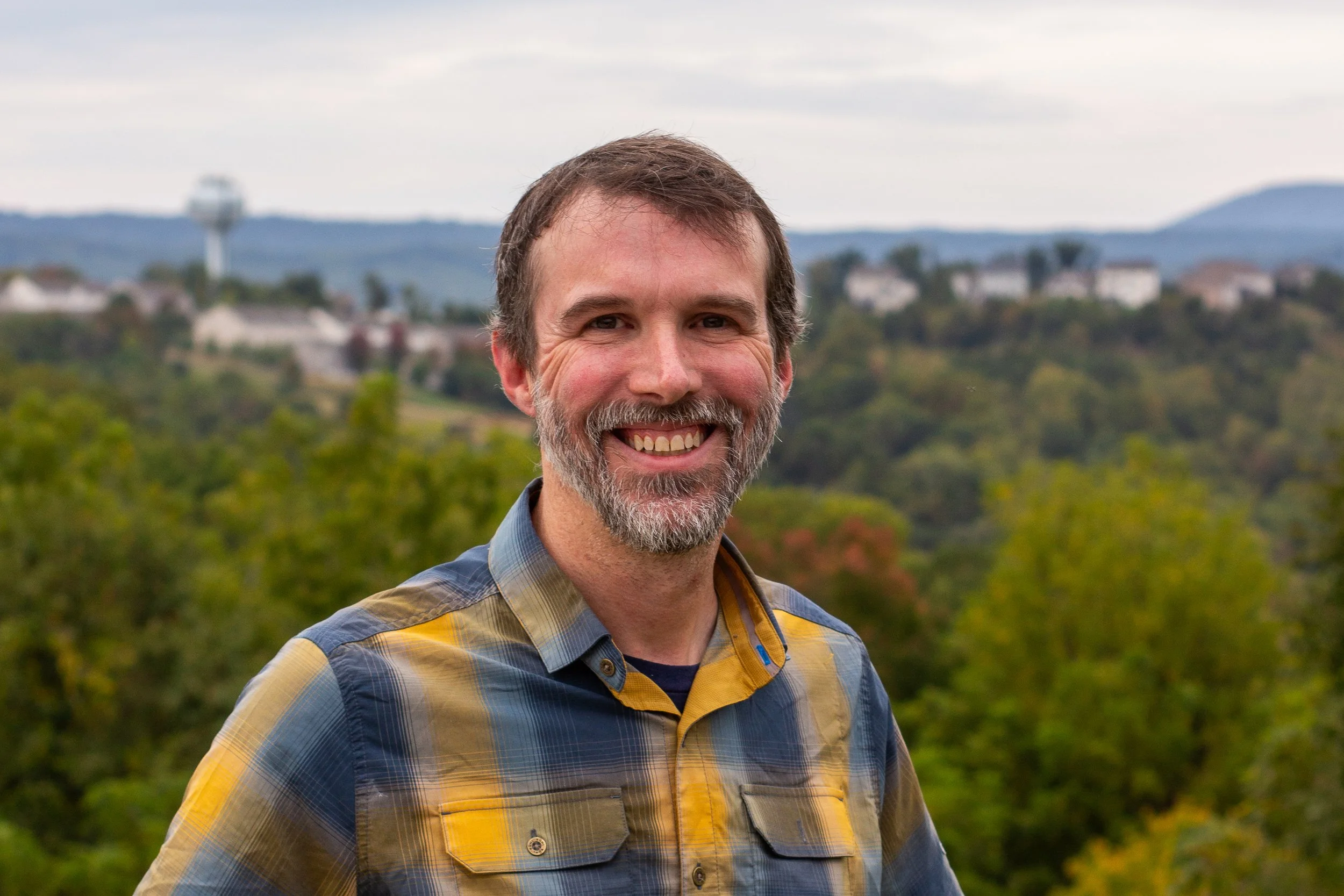A smiling middle-aged man with a beard and short brown hair outdoors, wearing a yellow and blue plaid shirt, with a scenic background of green trees, hills, and a cloudy sky.
