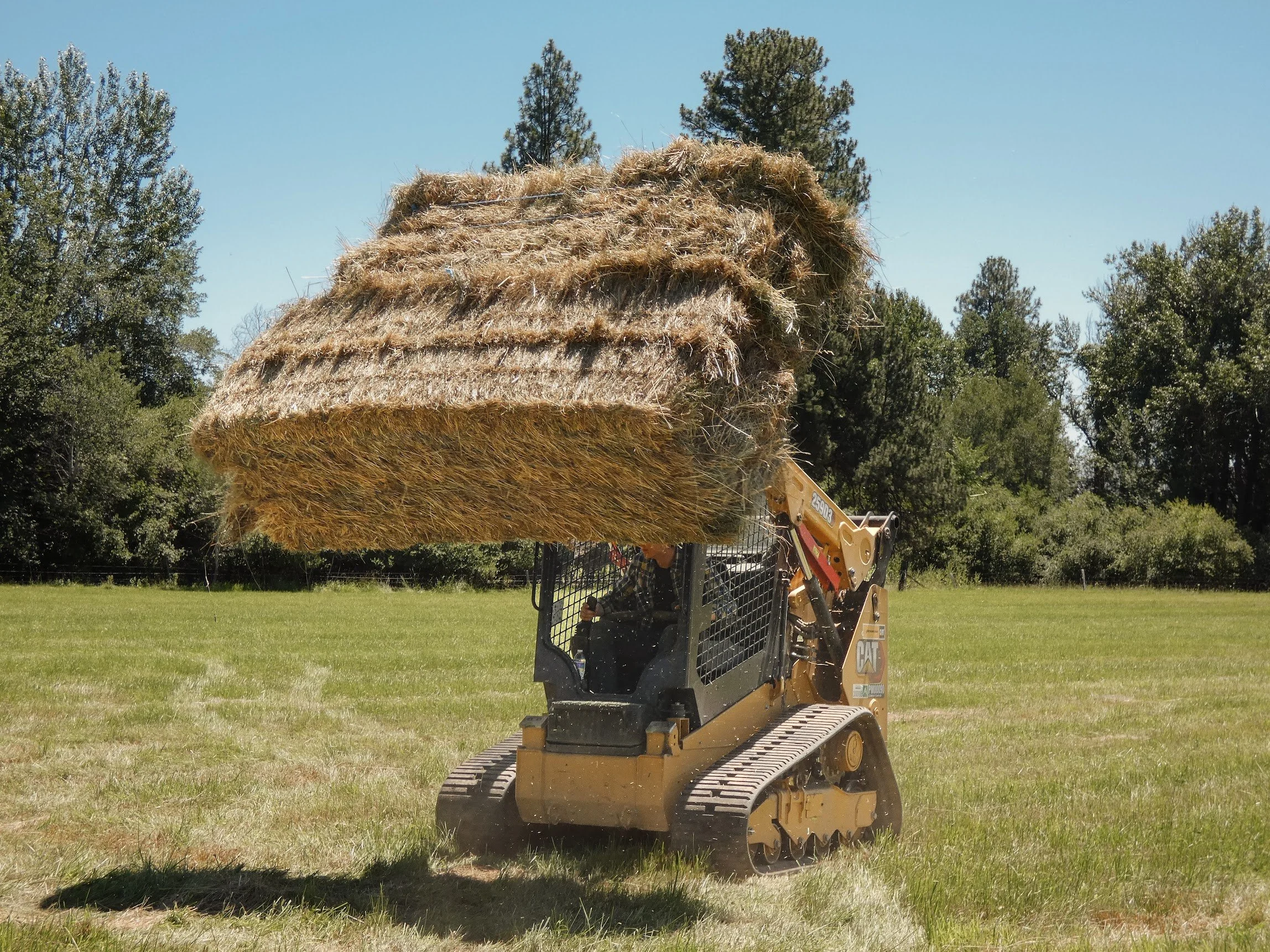 A small Caterpillar bulldozer lifting a large hay bale in a grassy field with trees in the background. Family Ranch located at Three Sons Ranch in Halfway Oregon.