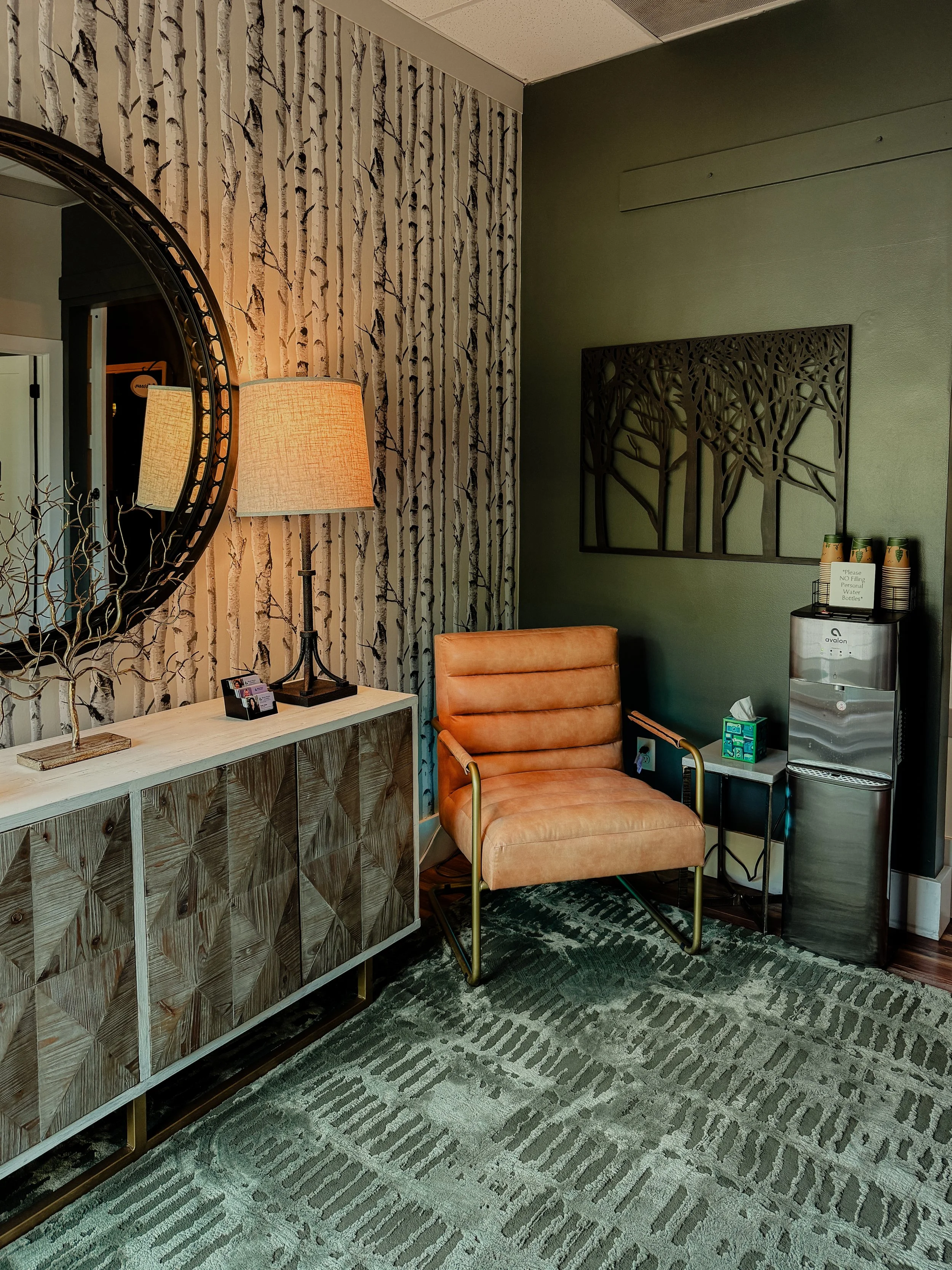 A cozy waiting area with a beige leather chair, a wooden sideboard with decorative pieces, a standing lamp with a beige shade, a round mirror on the wall, a green wall with tree artwork, and a water dispenser with cups.