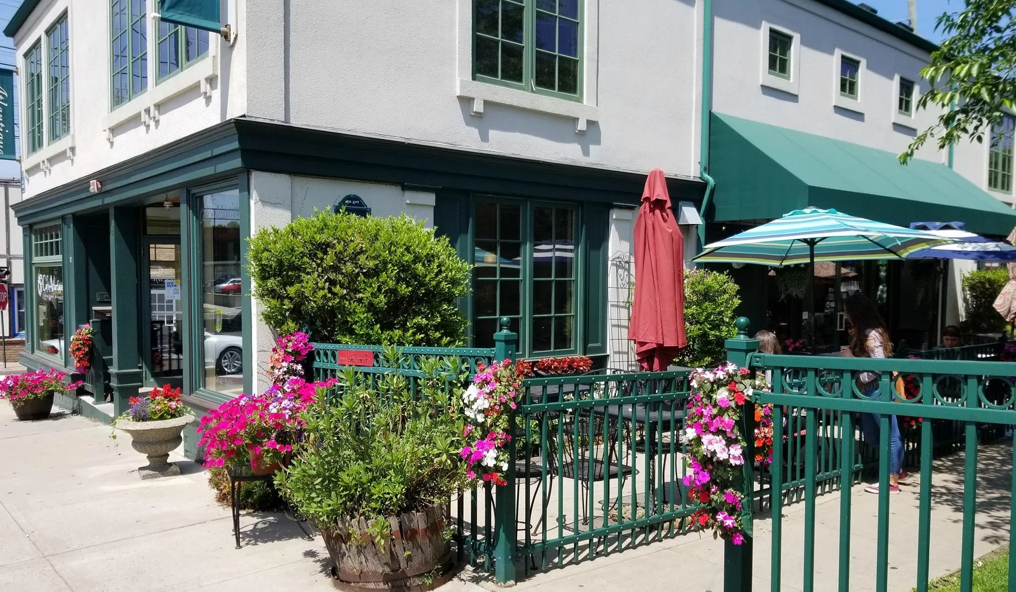 Exterior of a cafe or restaurant with outdoor seating, green railings, colorful flowers, and umbrellas on a sunny day.