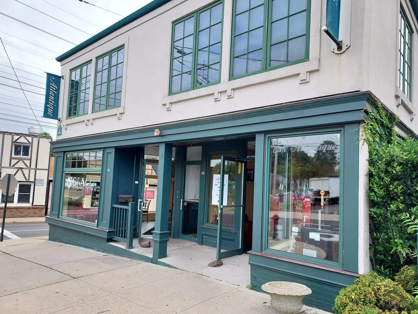 Exterior view of a two-story building with teal trim, housing a cafe named 'Cafe Antiques.' The storefront features large windows, a glass door, and a small porch with a railing. Signage visible on the building and window displays the cafe's name.