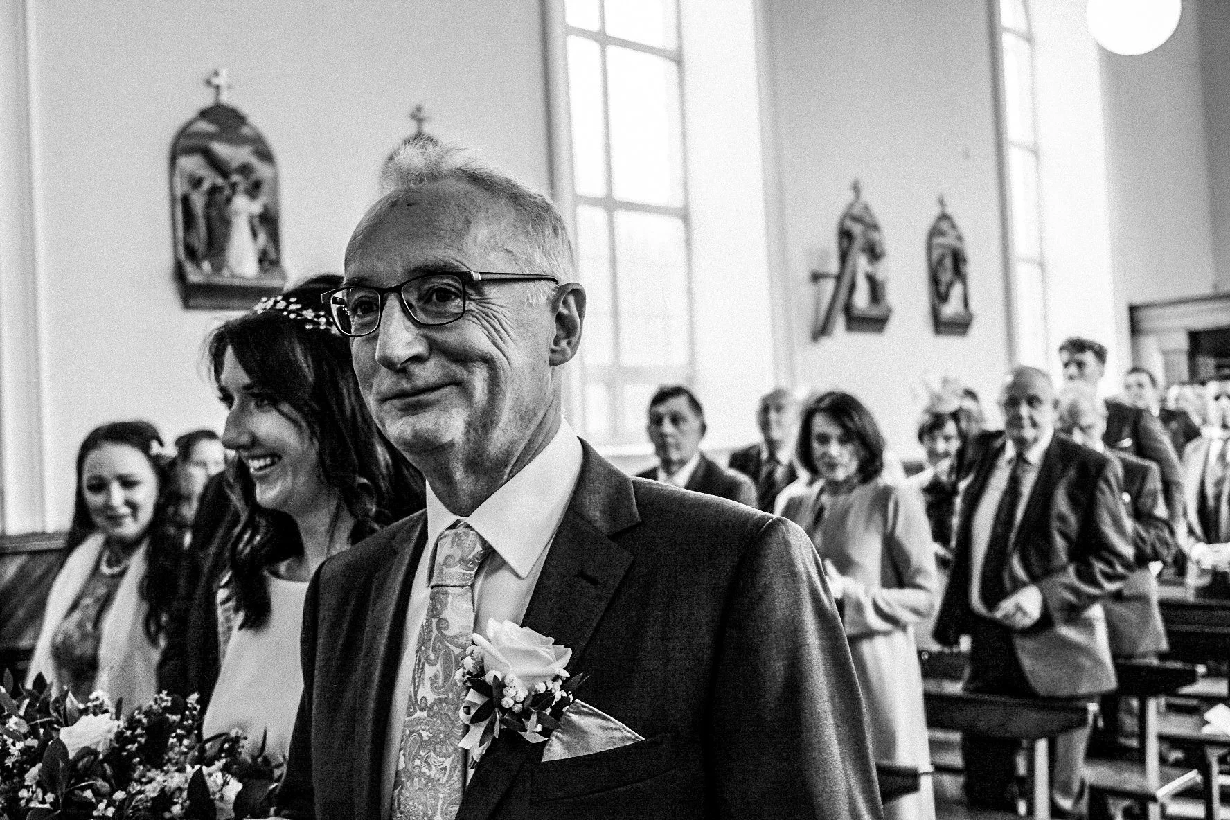 A black and white photo of a wedding ceremony inside a church, showing the groom smiling, wearing glasses, a suit, and a boutonniere, with the bridal party and guests in the background.