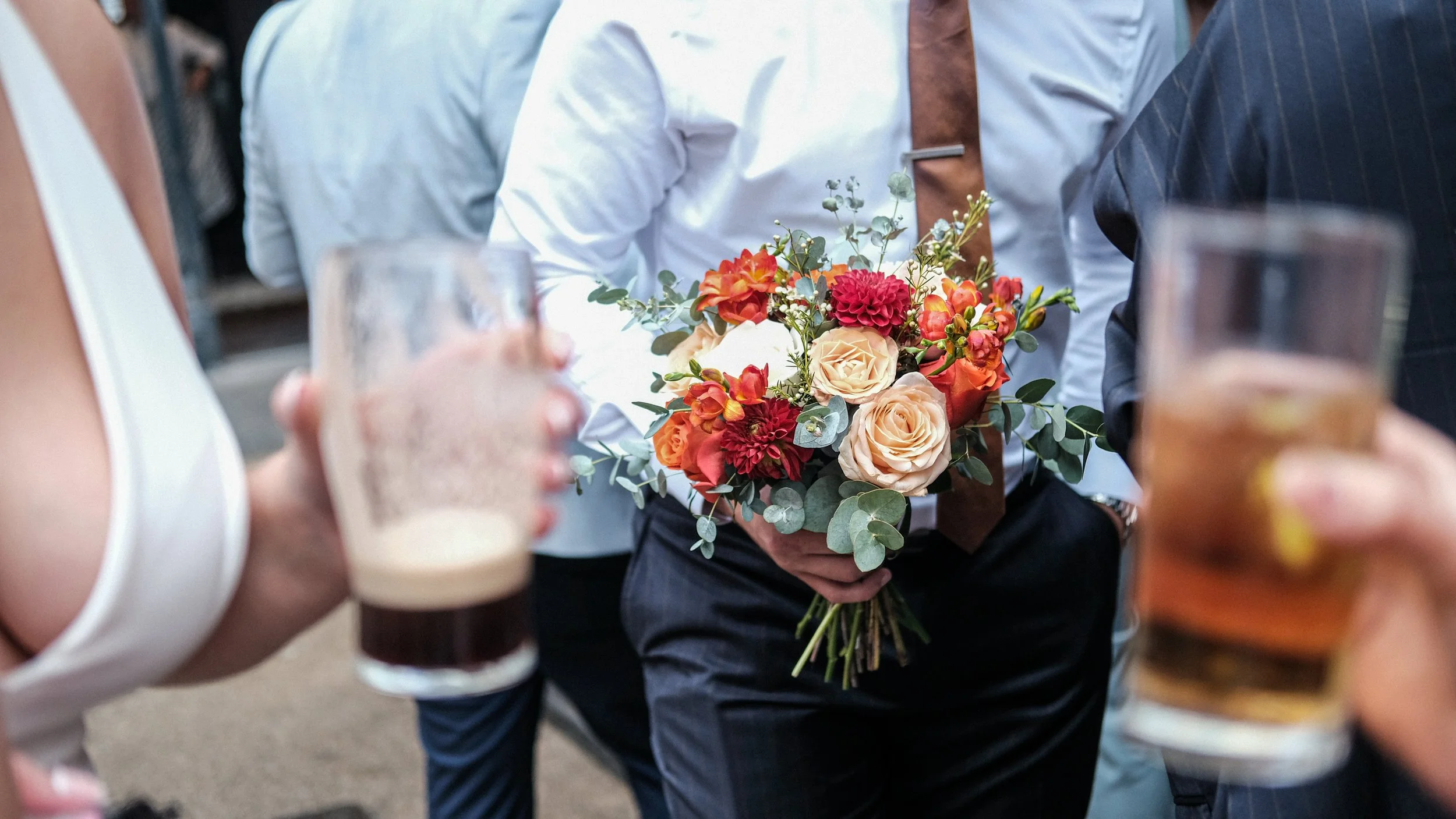 Person holding a bouquet of colorful roses, dahlias, and greenery at a social event, with others holding drinks nearby.