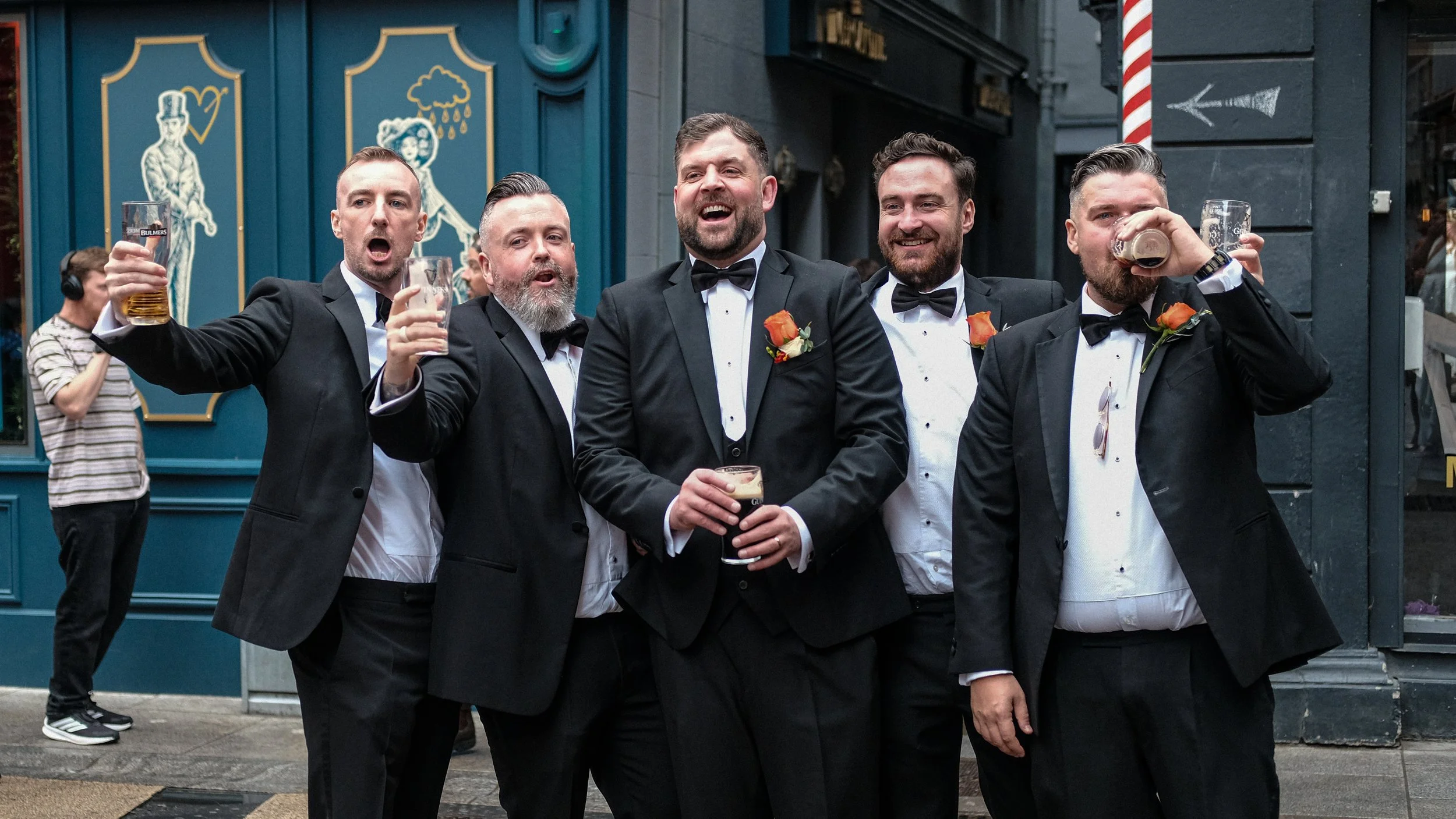 Group of five men in tuxedos celebrating outside a pub, holding drinks, with a woman in striped shirt in the background.