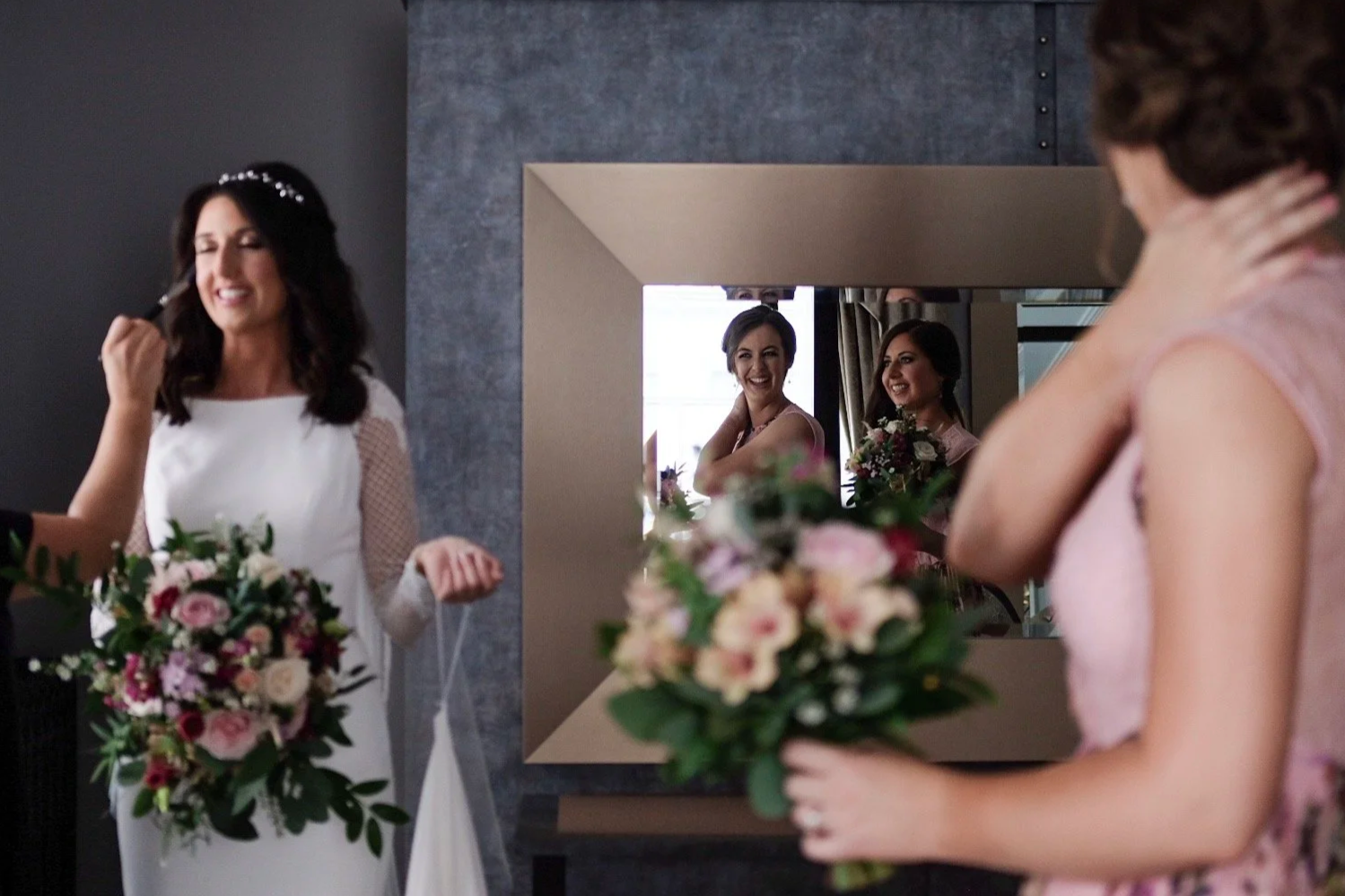 Bridesmaids and bride in front of a mirror at a wedding preparation, with bouquets of flowers, and friends smiling as they prepare.