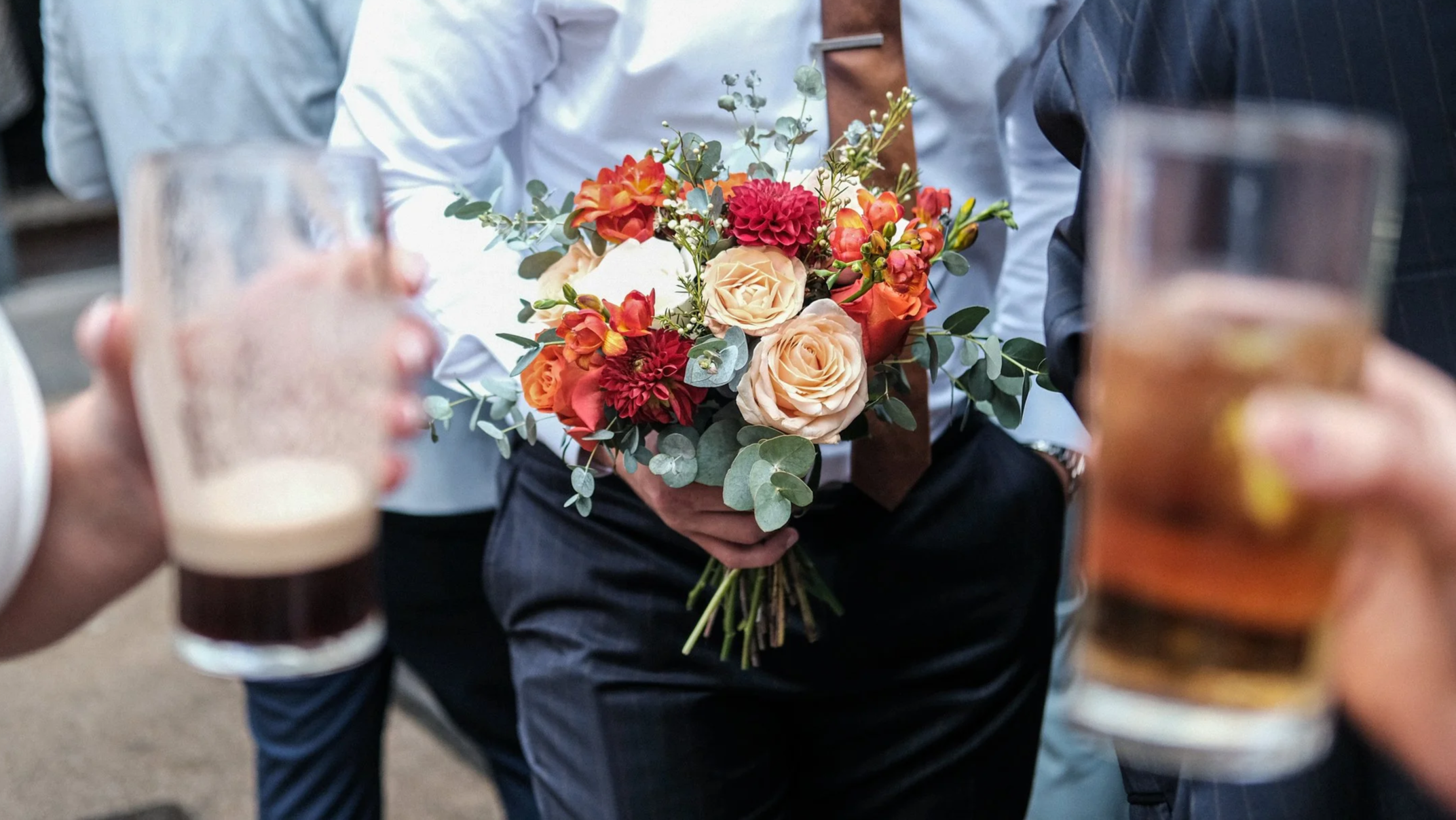 Person holding a bouquet of colorful roses, dahlias, and greenery at a social event, with others holding drinks nearby.