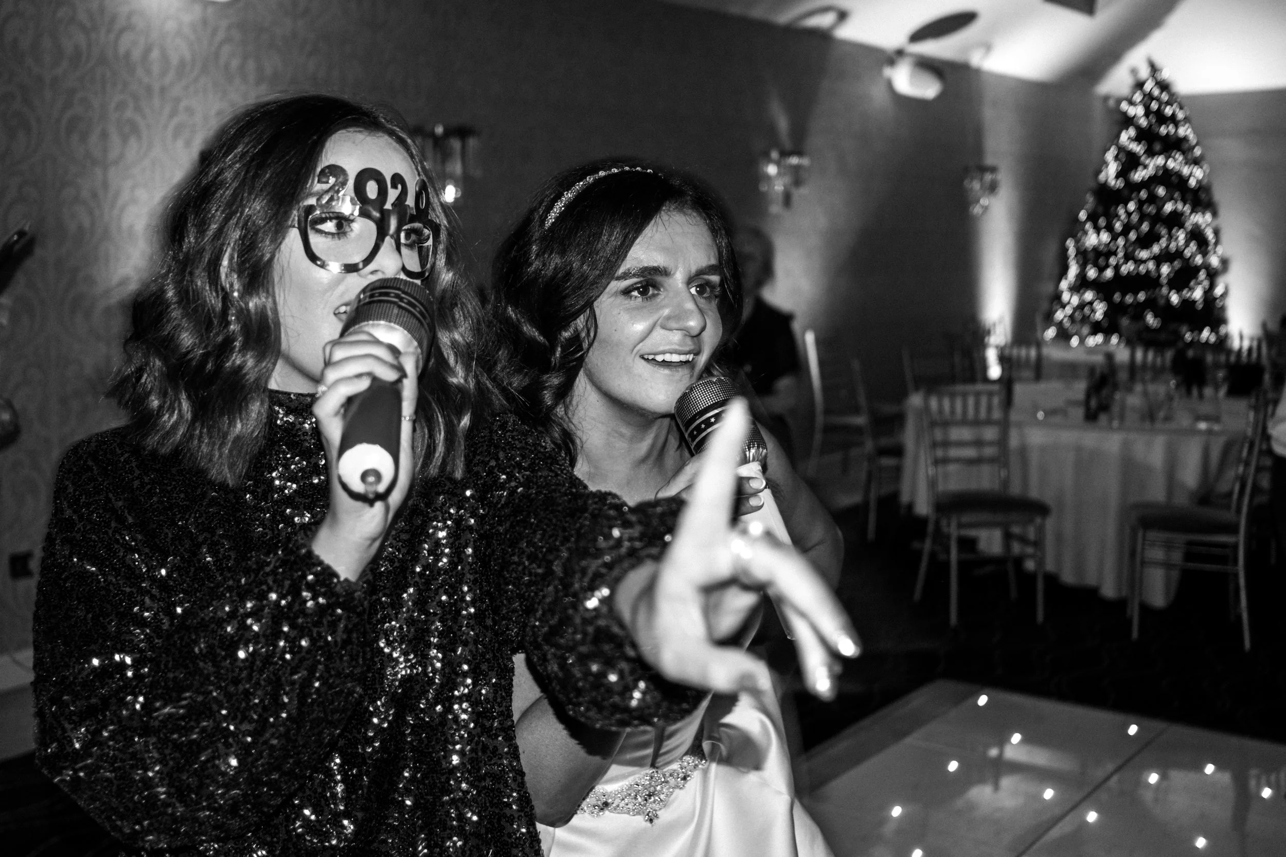 Two women at a celebration, with one wearing large glasses with '2023' and holding a microphone, the other reaching towards the camera, both smiling, with a Christmas tree and decorated tables in the background.