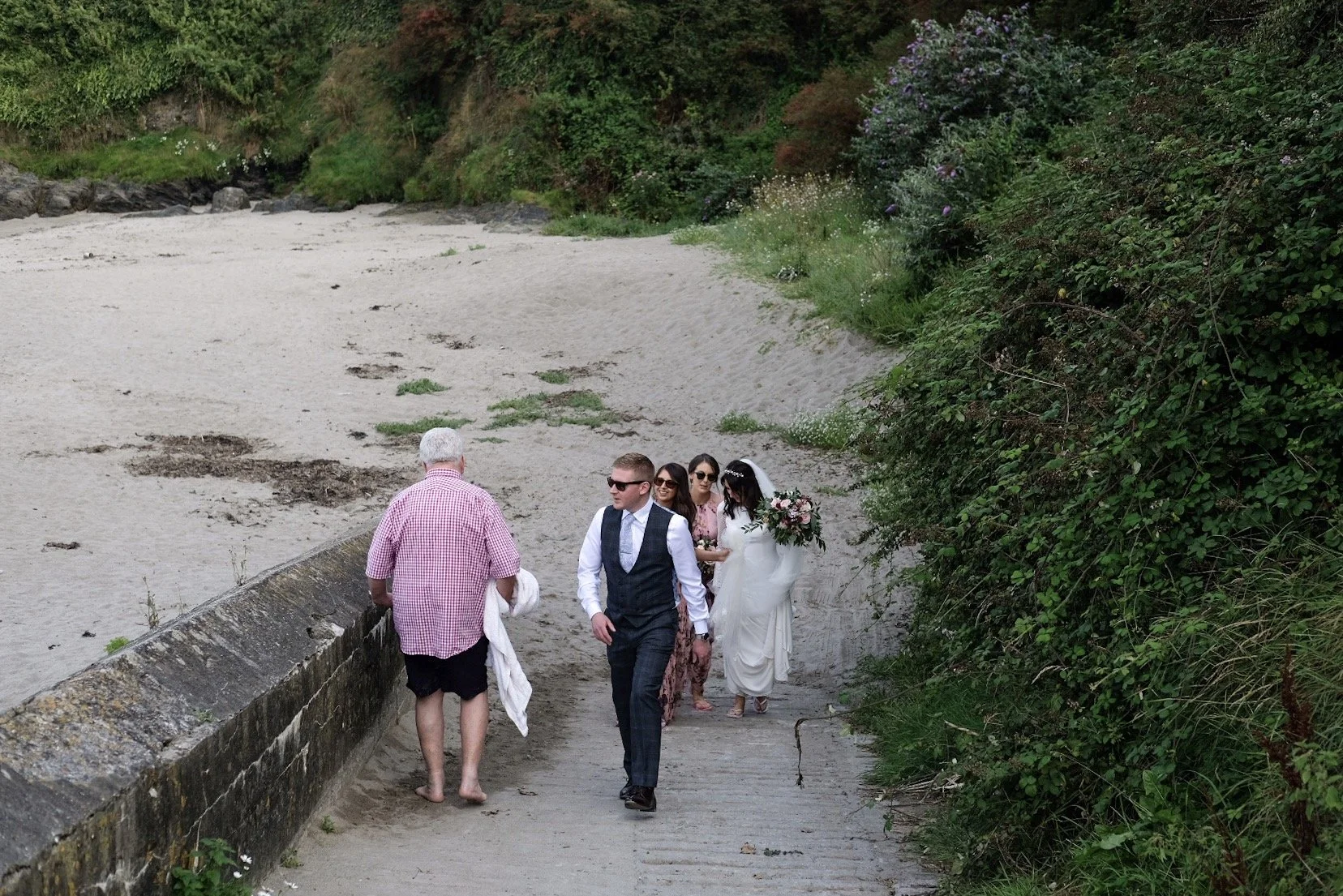 A group of people dressed in wedding attire walking on a sandy pathway along a beach, with lush green bushes and trees on the right side.