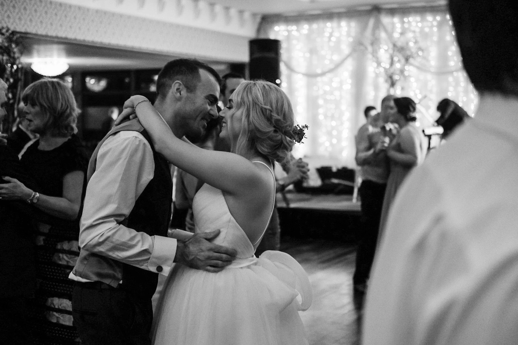 A black and white photo of a couple dancing at a wedding reception, with the bride and groom smiling and close to each other. Other guests are visible in the background, some dancing and others watching.