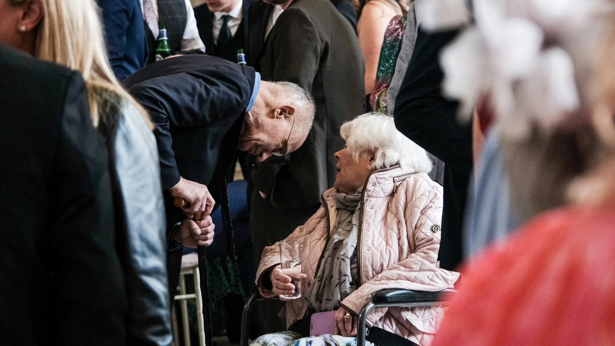 An elderly woman with white hair seated in a wheelchair, holding a glass of water, engaged in conversation with a man leaning over her at a social gathering or event surrounded by many people.