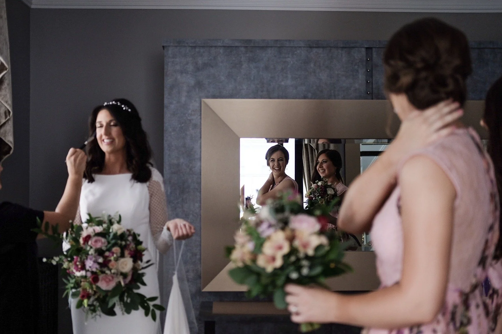Bridesmaids and bride in front of a mirror at a wedding preparation, with bouquets of flowers, and friends smiling as they prepare.