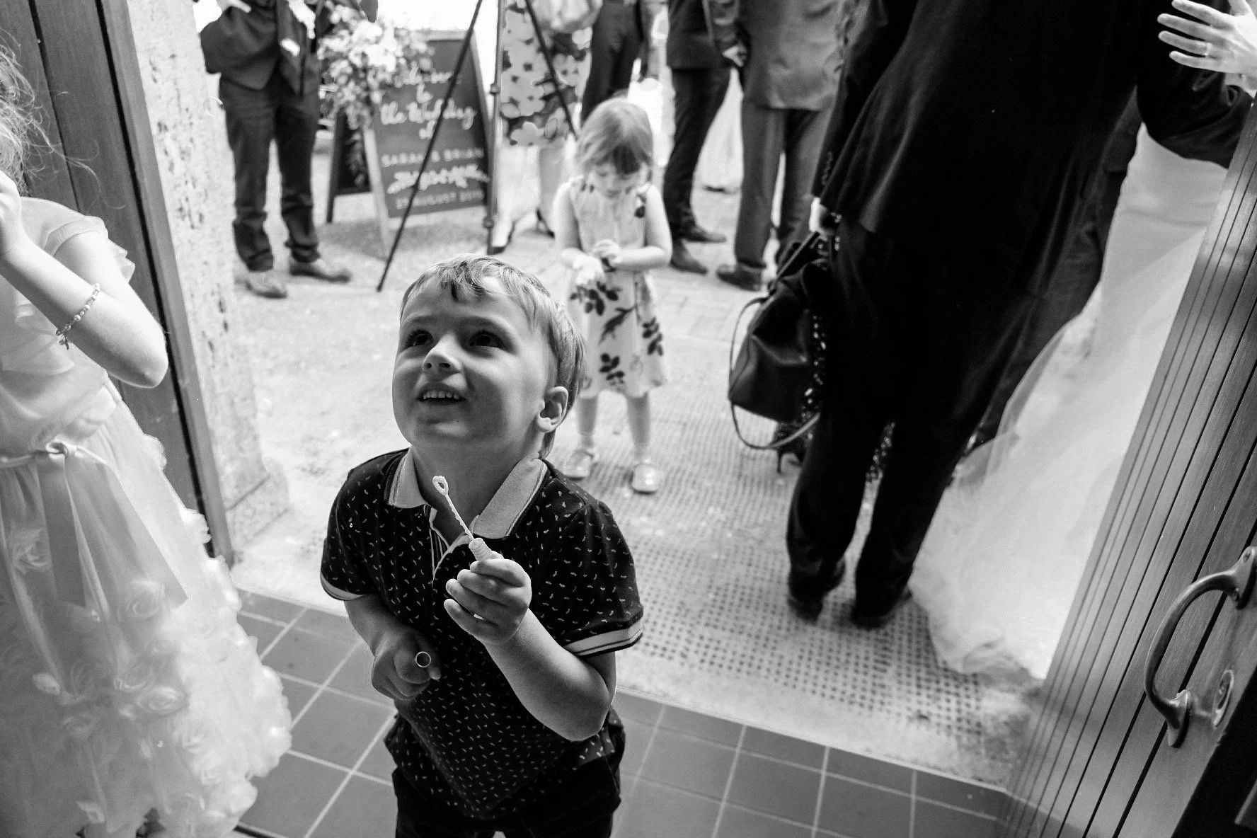 A young boy smiling and looking upwards, standing in an indoor space near the entrance, holding a small balloon or string. Behind him, two young girls are outside, one looking down and the other looking — possibly at a phone. Several adults are stand
