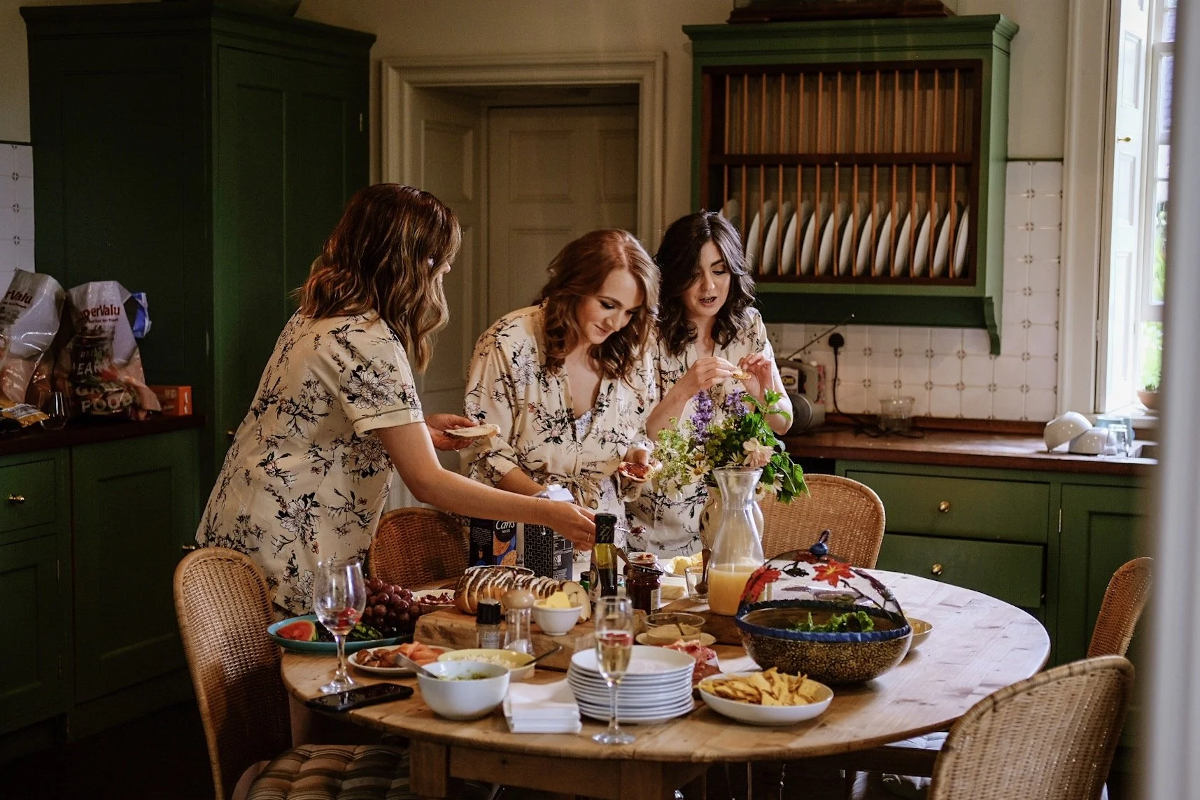Three women in matching floral pajamas preparing food and drinks at a cluttered kitchen table, with various dishes, bowls, snacks, and a pitcher of orange juice, in a cozy kitchen with green cabinets and a window.
