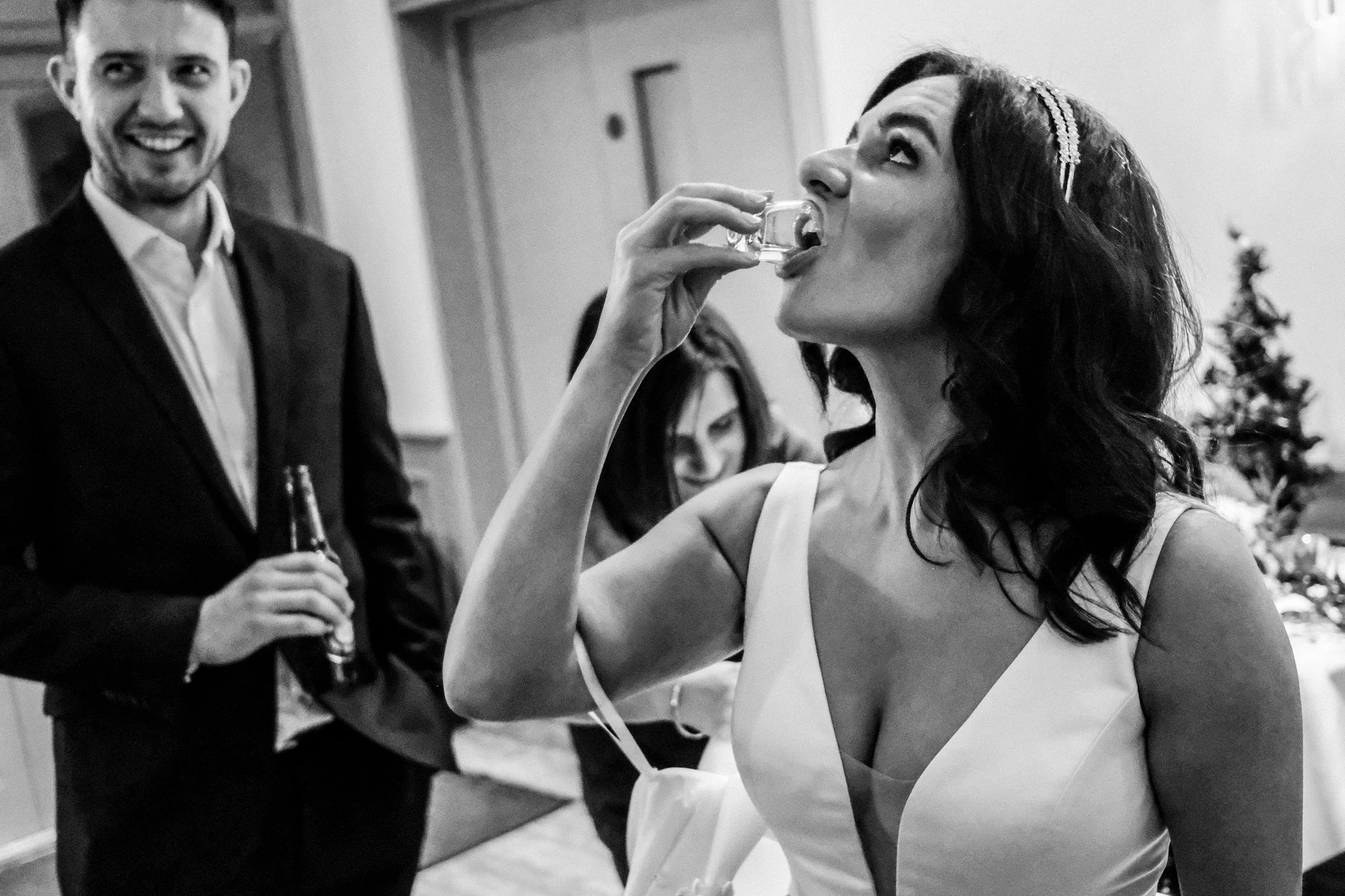 A woman in a sleeveless dress drinks from a small glass at a social gathering, with a man in a suit holding a drink smiling in the background.