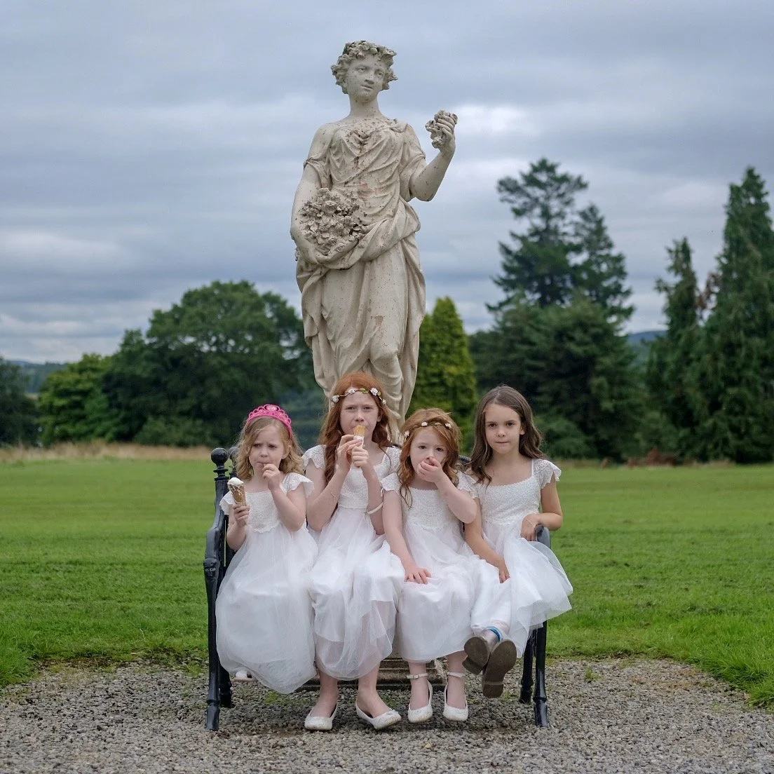Four young girls wearing white dresses sitting on a bench in front of a large stone statue of a woman holding flowers, outdoors on cloudy day with trees in the background.
