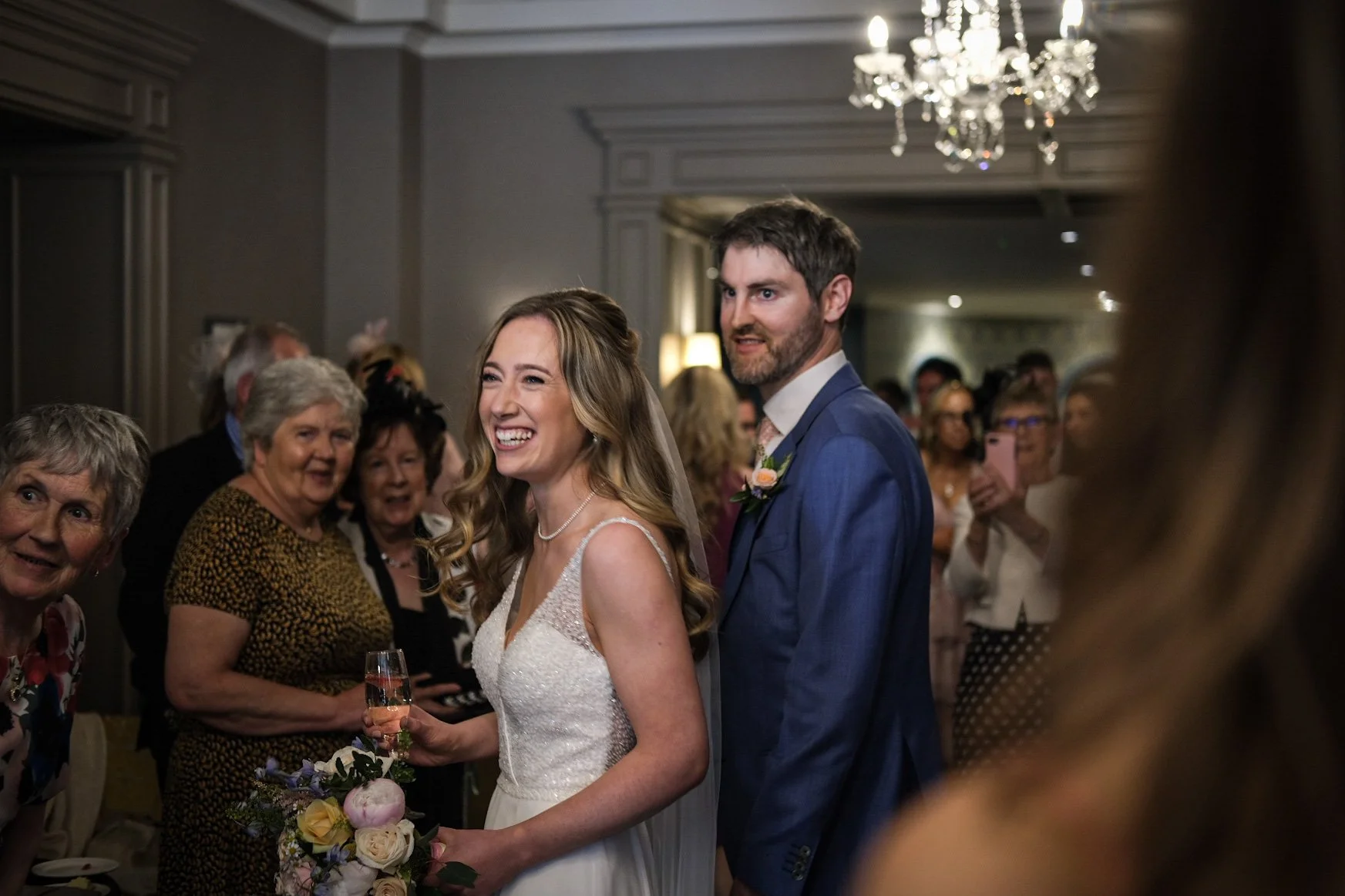 Smiling bride in white wedding dress holding a bouquet of flowers and standing next to groom in blue suit during wedding reception with guests in background.