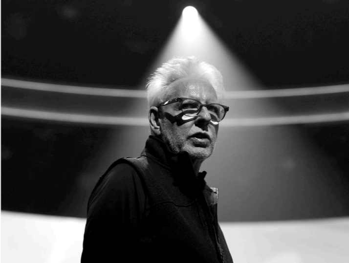 A black and white photo of a man with white hair and glasses, standing under a spotlight in a dark setting, possibly on a stage or in a studio.