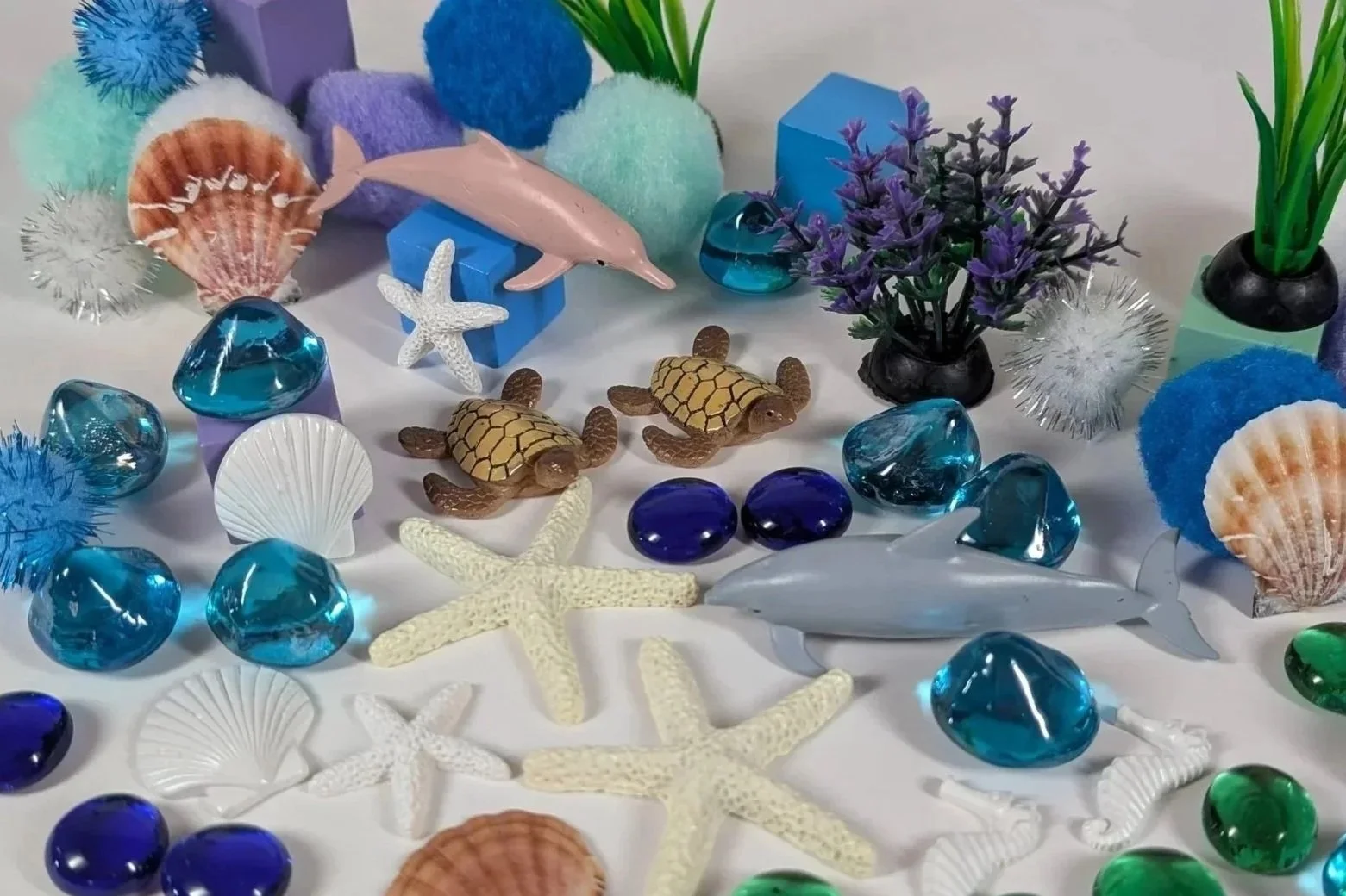 Play Sparks Ocean Spark Box open with loose parts including dolphin and sea turtle figurines, starfish, seashells, a seahorse, aquatic plants, blue and green gem stones, and colorful pom poms — open-ended ocean themed toy gift for kids ages 4 to 8