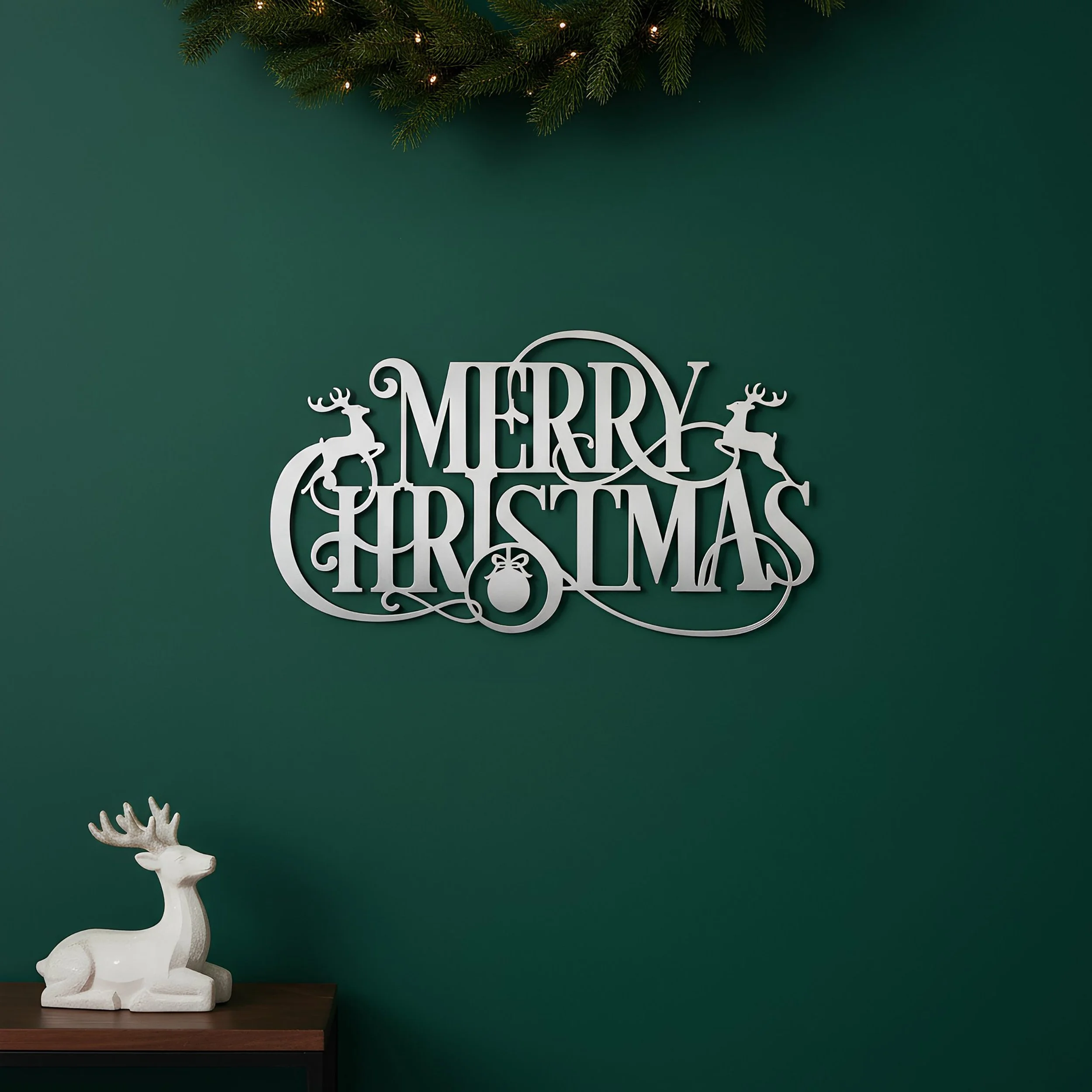 White metal sign with the text 'Merry Christmas' printed in an elegant style, with reindeer and ornament designs against a dark green wall, with a white reindeer figurine on the table below.
