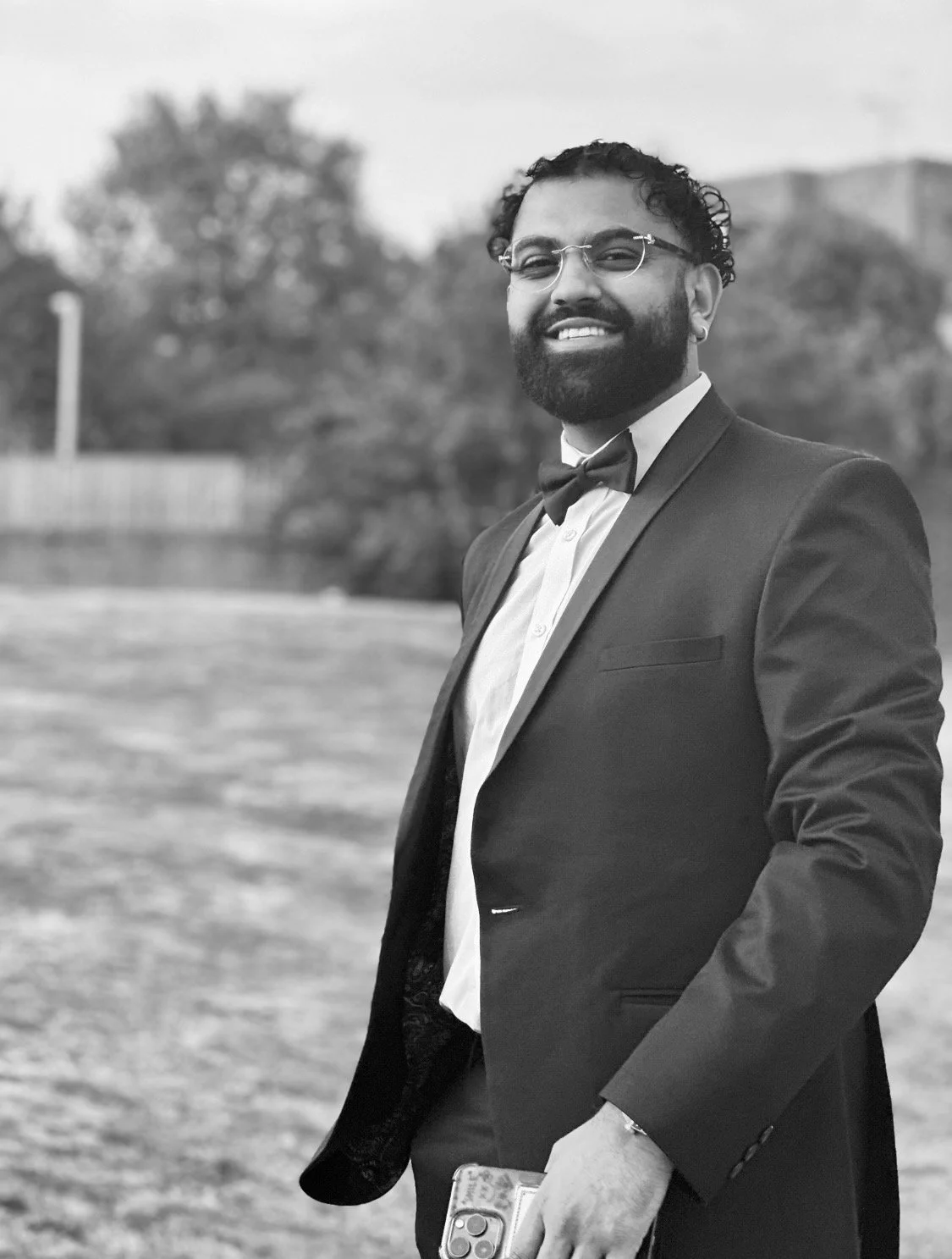 A man dressed in a tuxedo with a bow tie, glasses, and a beard, standing outdoors and smiling, holding a smartphone in his hand | Dry Eye