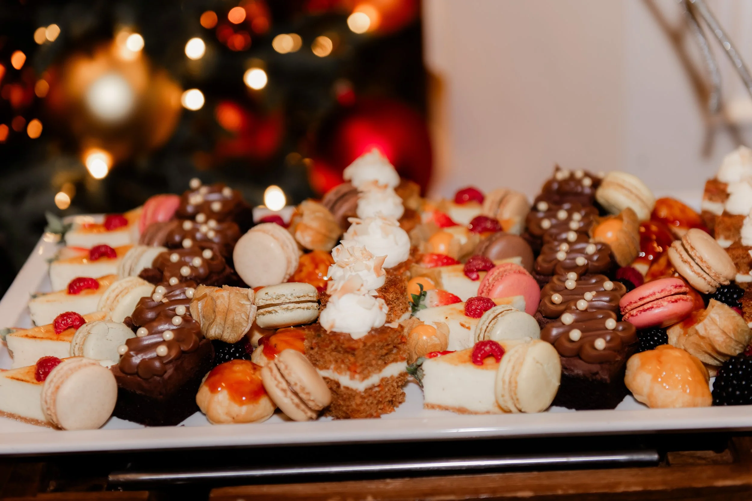 A tray of assorted desserts including macarons, chocolates, and cakes, with a blurred Christmas tree and ornaments in the background.