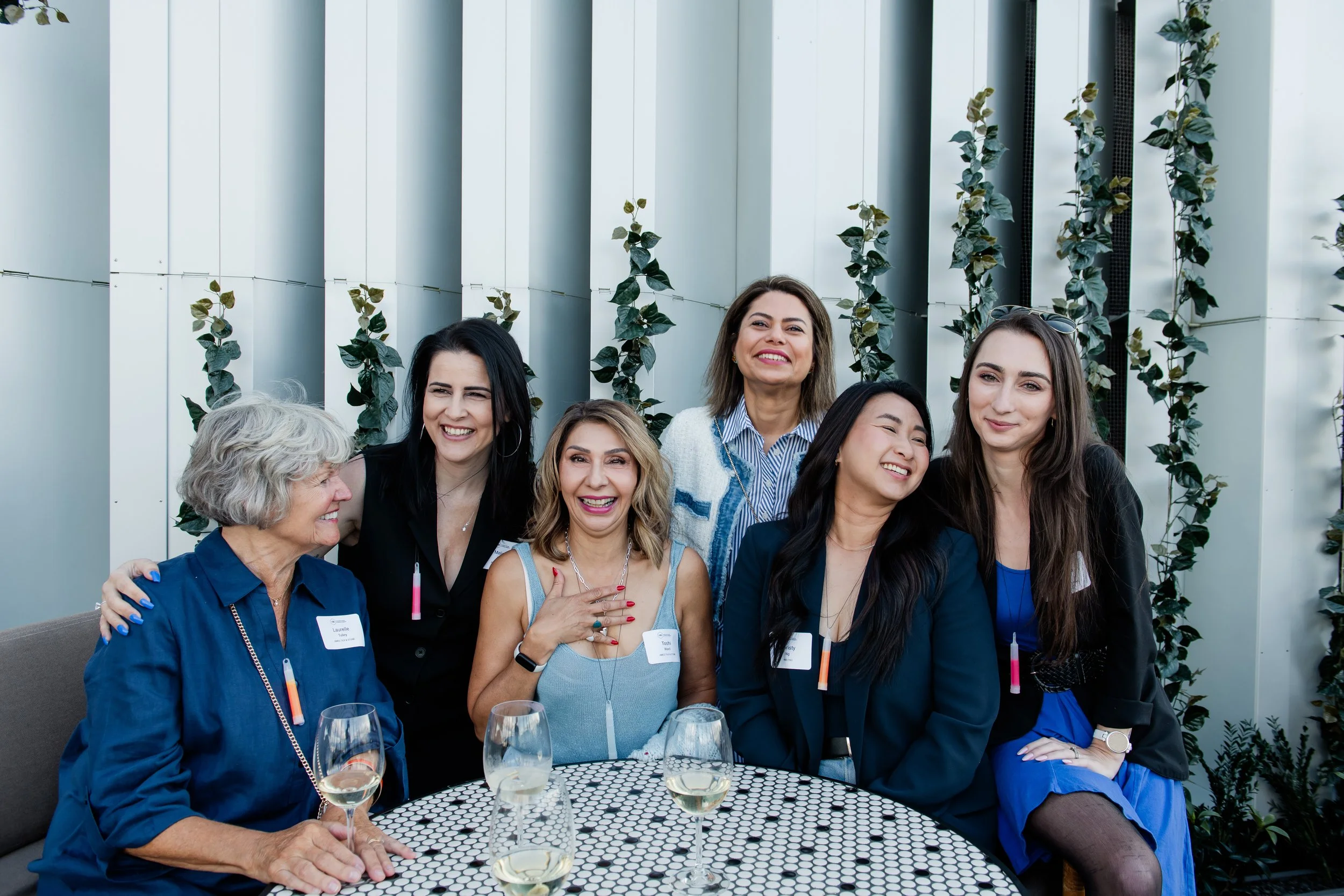Group of seven diverse women at a social gathering, sitting and standing around a table with wine glasses, smiling and enjoying each other's company outdoors.