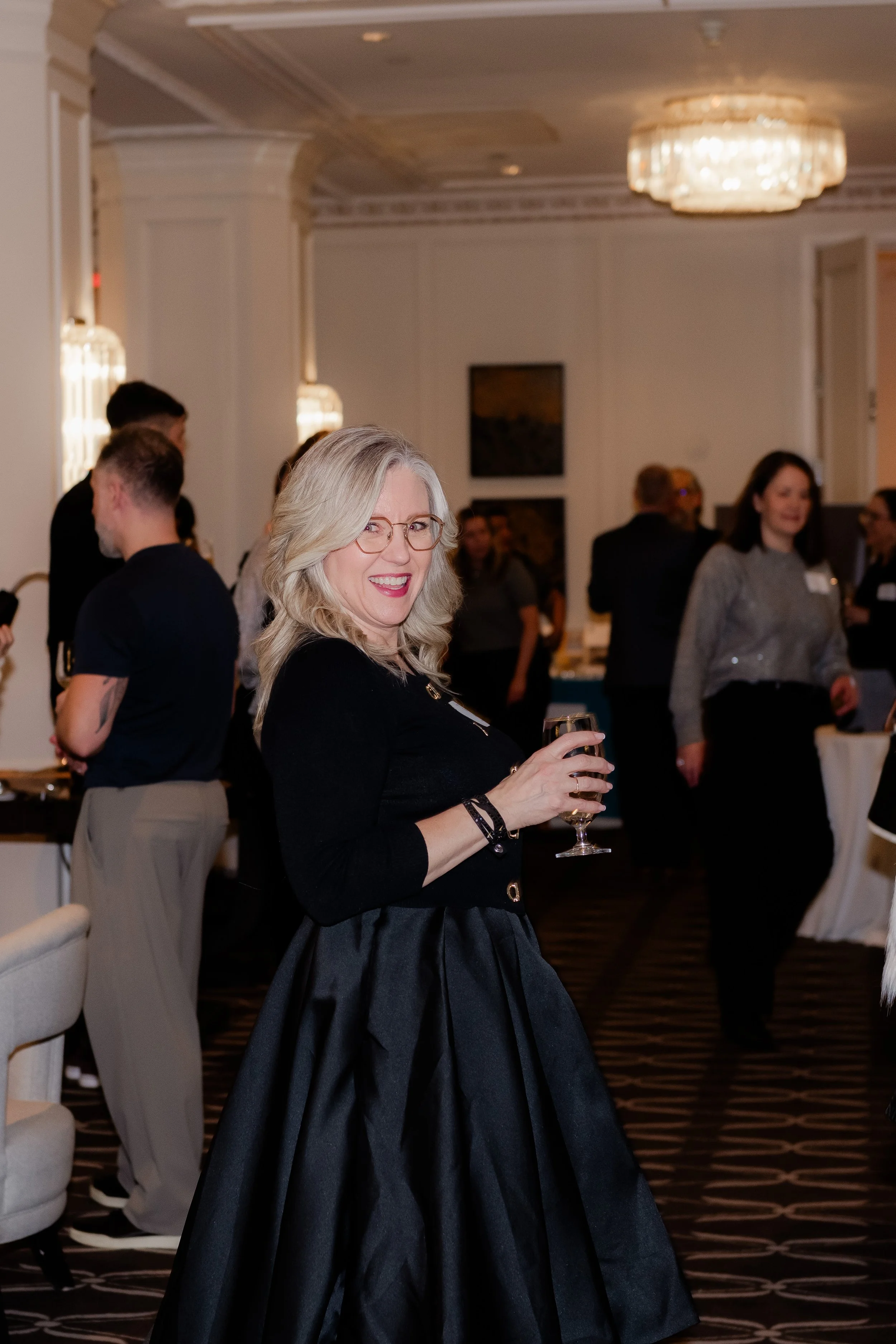 A smiling woman with glasses and blonde hair holding a wine glass at a social event in an elegant ballroom with chandeliers and other guests in the background.