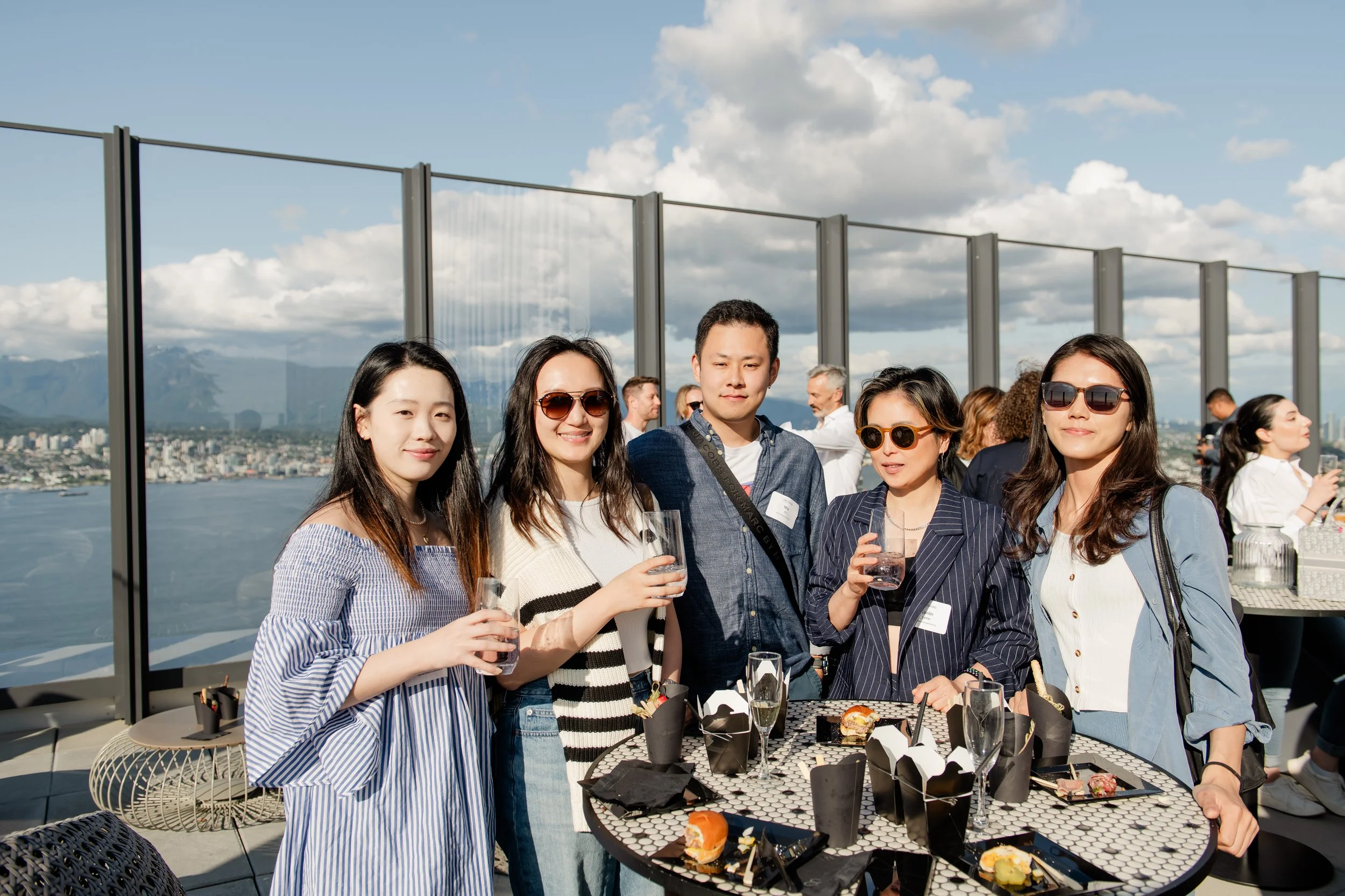 Group of five young adults standing on a rooftop with a city and river view, holding drinks, with other people in the background on a sunny day.