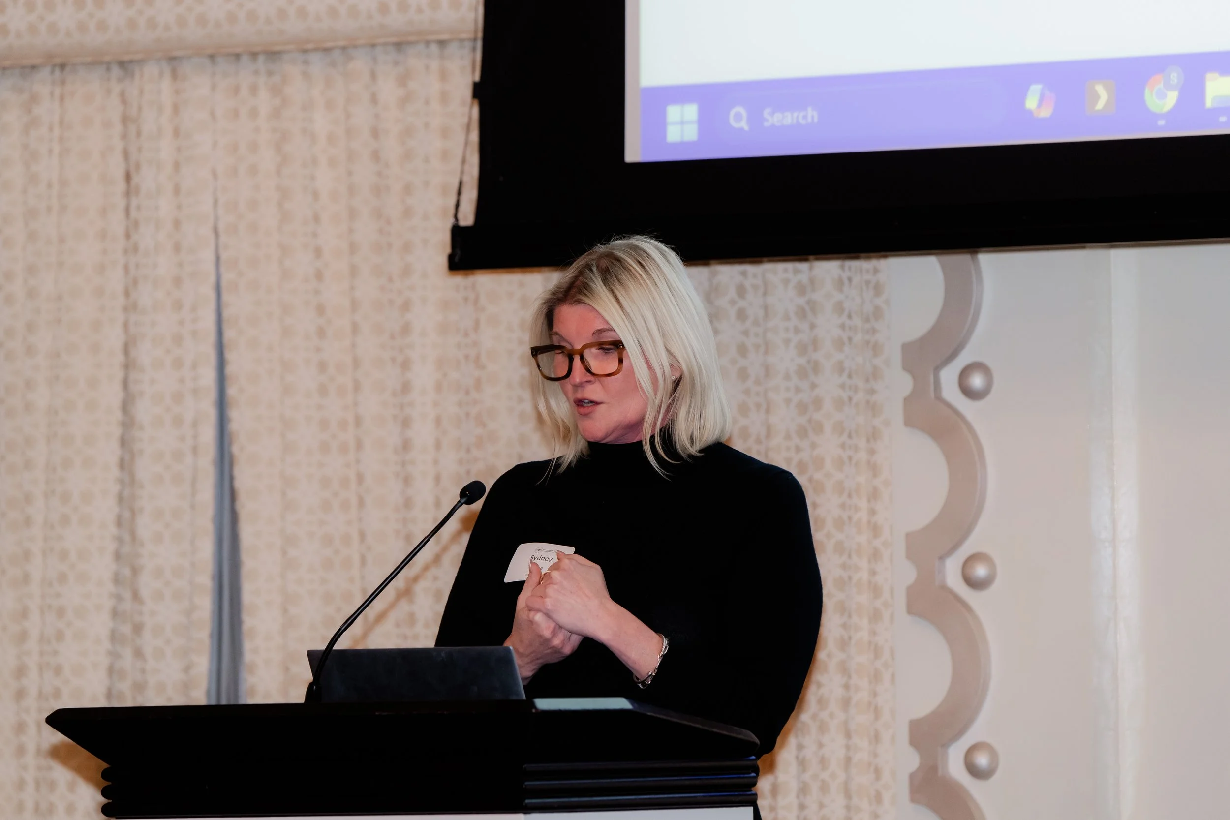 A woman with blonde hair, glasses, and a black top speaking at a podium with a microphone, with a large screen behind her showing part of a computer desktop.