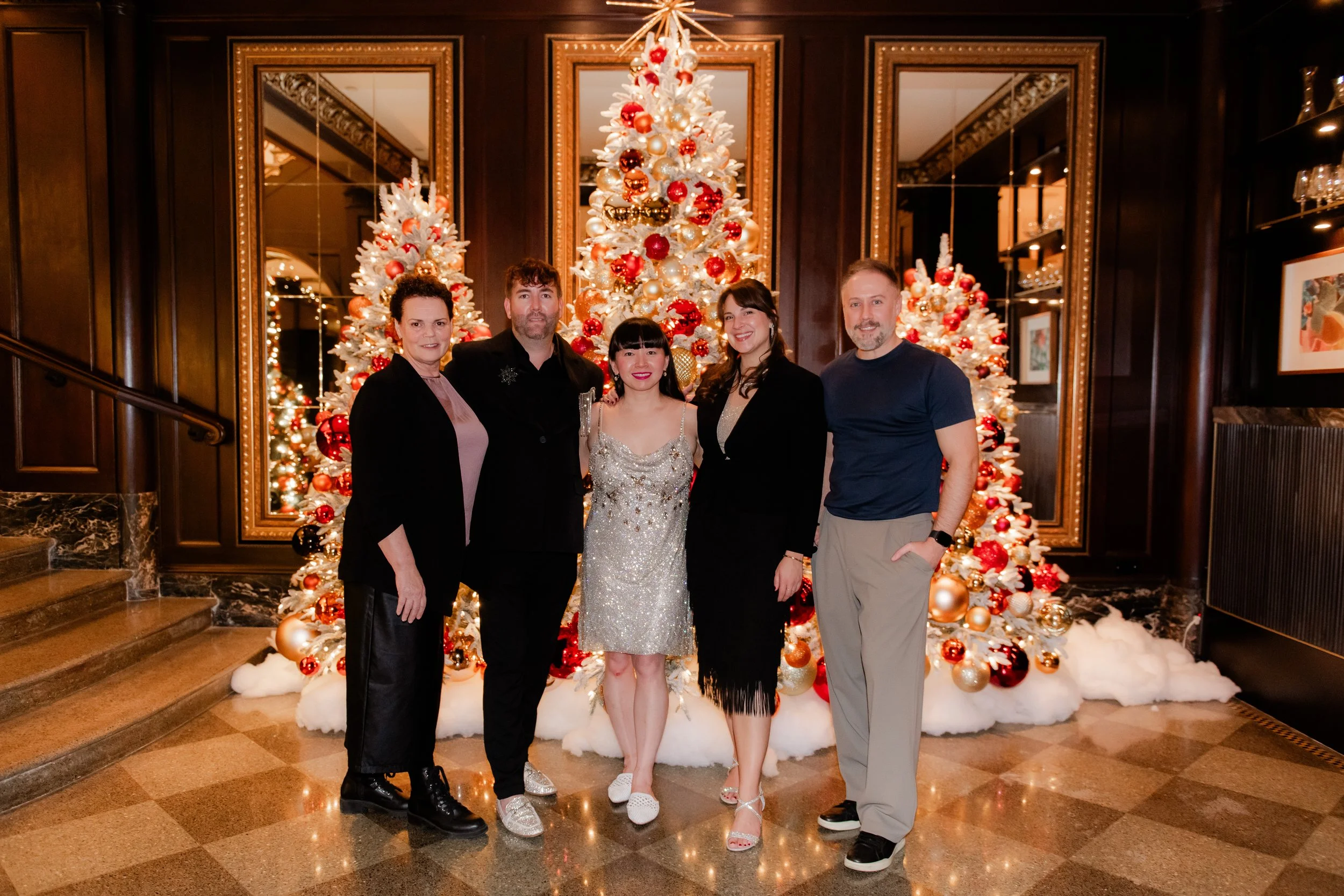 A group of five people posing in front of a decorated Christmas tree inside a festive room with wood-paneled walls and mirrored panels.